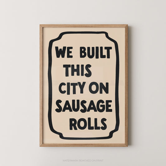 Framed wall art print with the phrase “WE BUILT THIS CITY ON SAUSAGE ROLLS” in bold black text inside a cream rectangle, set in a light natural wood frame against a light gray wall.