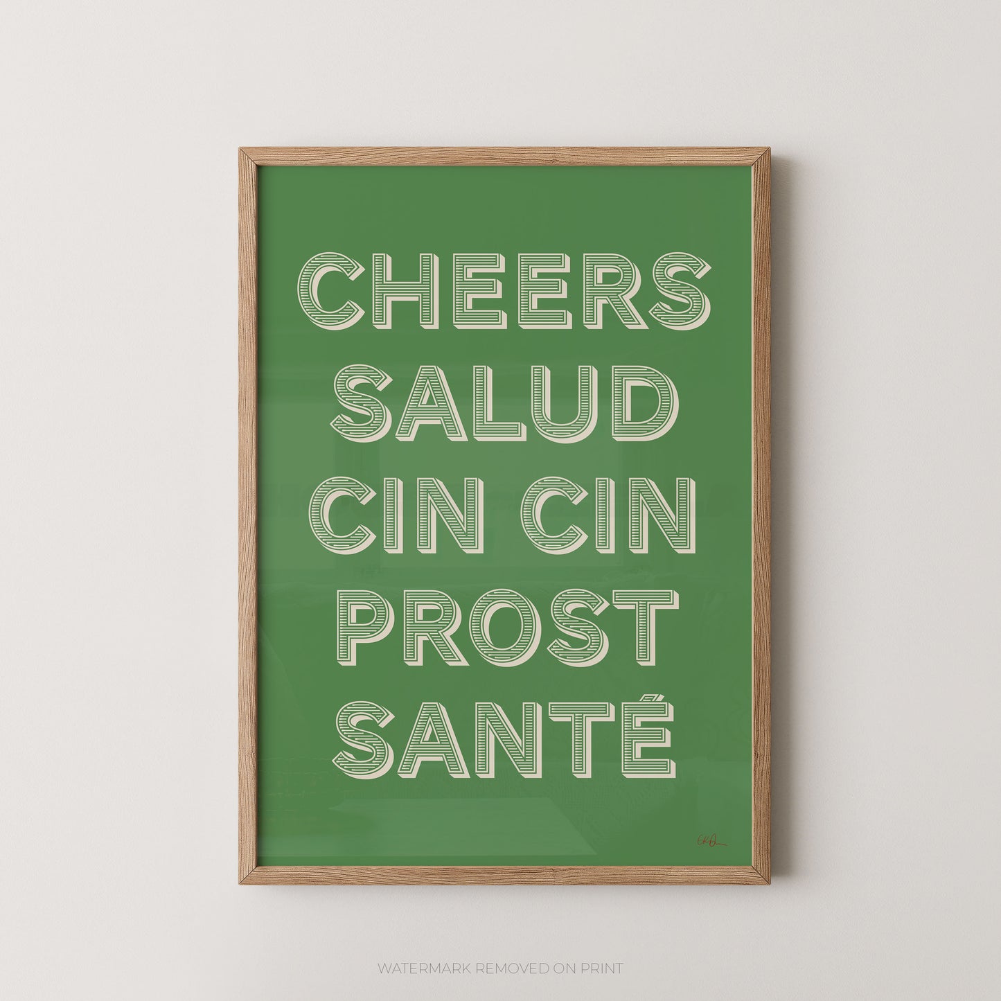 A framed art print featuring the text 'CHEERS SALUD CIN CIN PROST SANTÉ' in raised white letters on a solid green background, enclosed in a light wood frame against a neutral light gray wall.