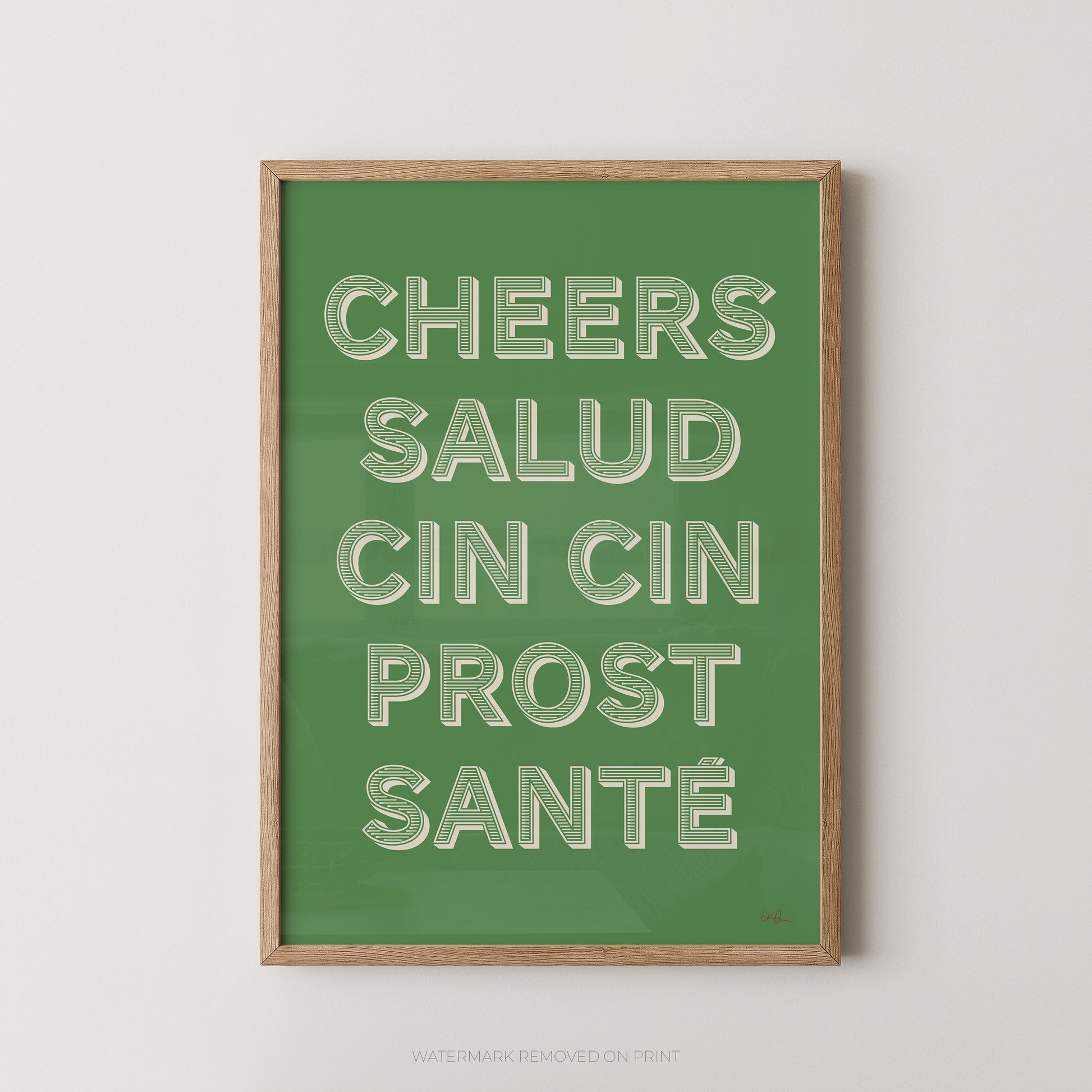 A framed art print featuring the text 'CHEERS SALUD CIN CIN PROST SANTÉ' in raised white letters on a solid green background, enclosed in a light wood frame against a neutral light gray wall.