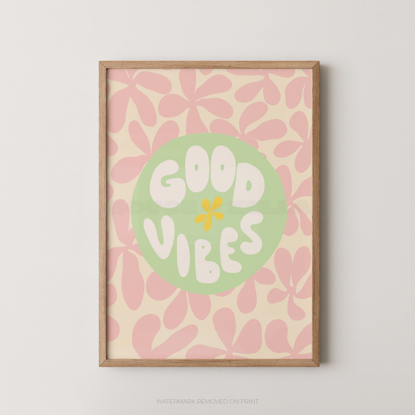A framed art print featuring the words 'GOOD VIBES' in white retro lettering inside a light green circle, set against a pink and cream floral pattern. The print is housed in a light natural wood frame and is displayed on a neutral light gray wall.