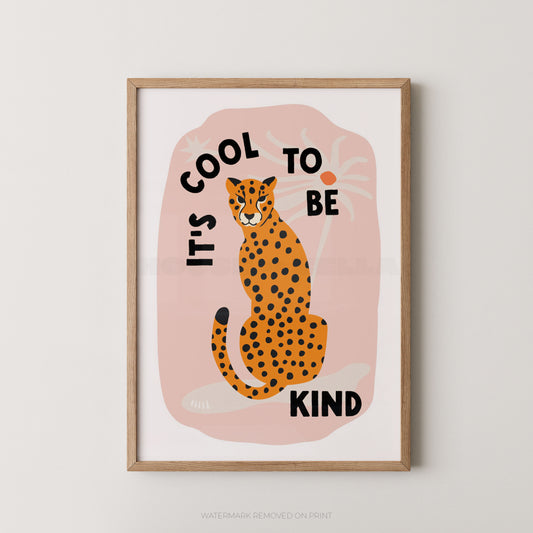 A framed art print featuring an orange and black spotted cheetah sitting on a pink background with the text 'IT'S COOL TO BE KIND' in black lettering, displayed on a light gray wall.