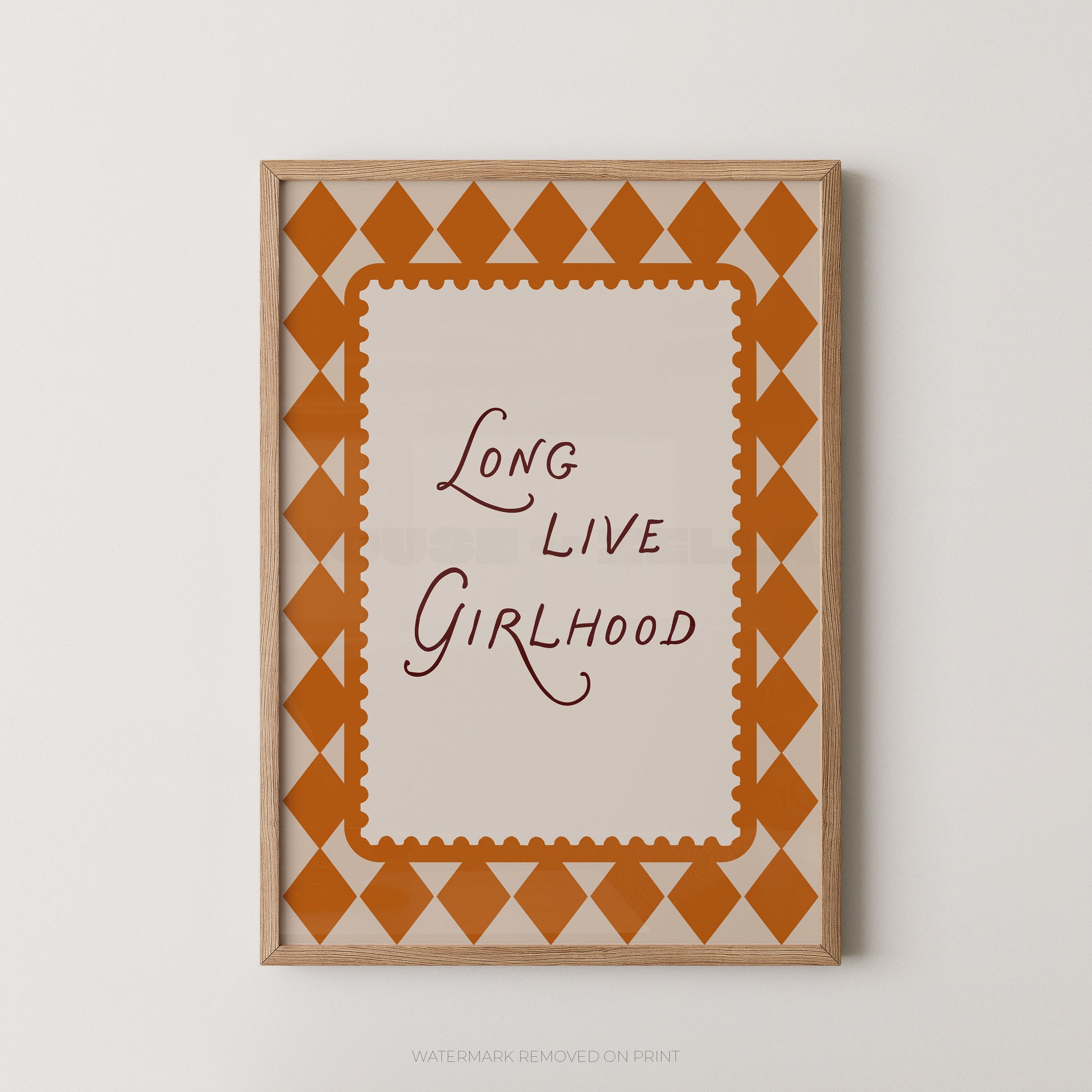 Framed wall art print with the phrase 'Long Live Girlhood' in dark script inside a cream center panel, surrounded by an orange diamond pattern and a light wood frame, shown against a light neutral wall.