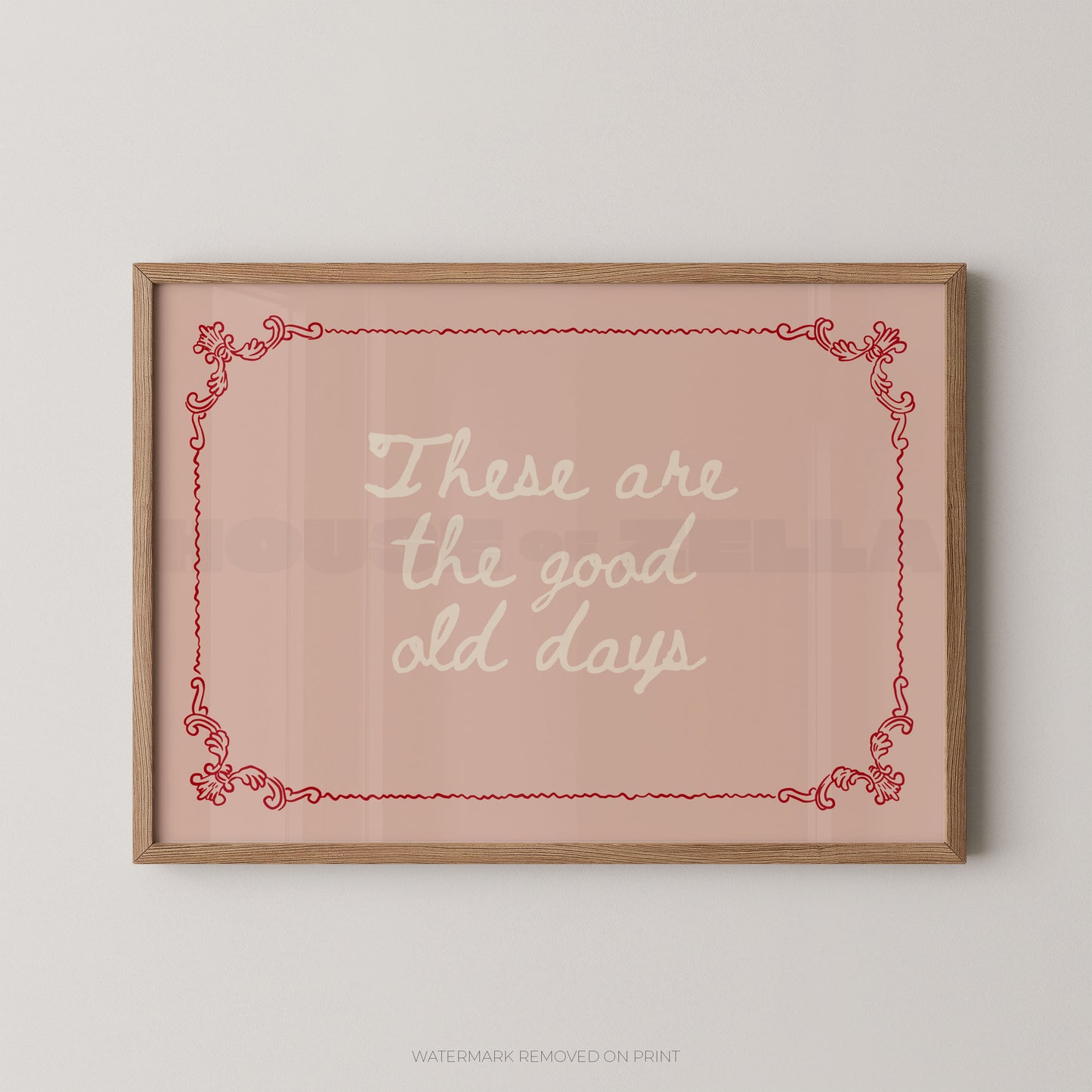 Framed wall art print with the phrase “These are the good old days” in white script on a dusty pink background, bordered by a red decorative frame and a light wood frame, shown hanging on a light gray wall.