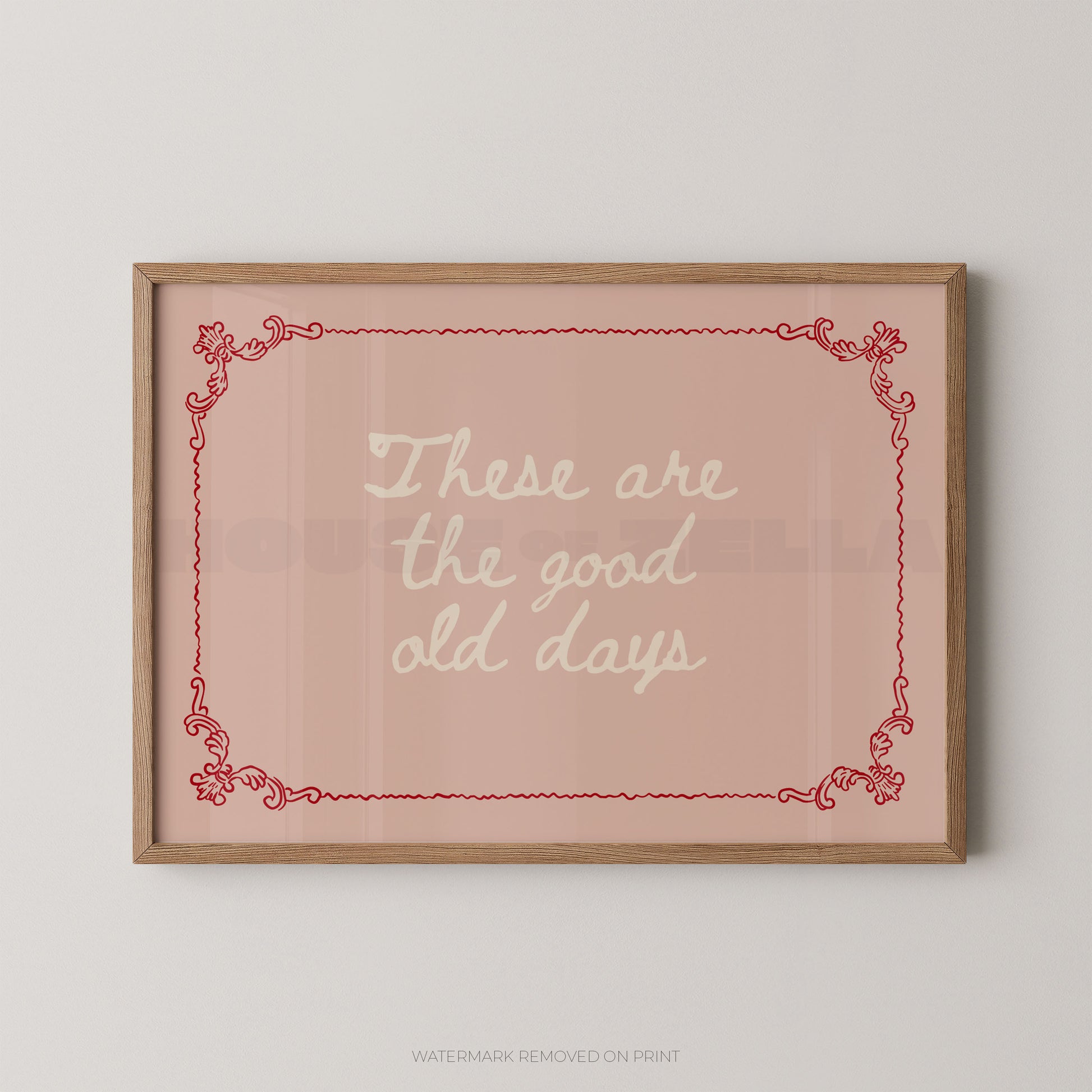 Framed wall art print with the phrase “These are the good old days” in white script on a dusty pink background, bordered by a red decorative frame and a light wood frame, shown hanging on a light gray wall.