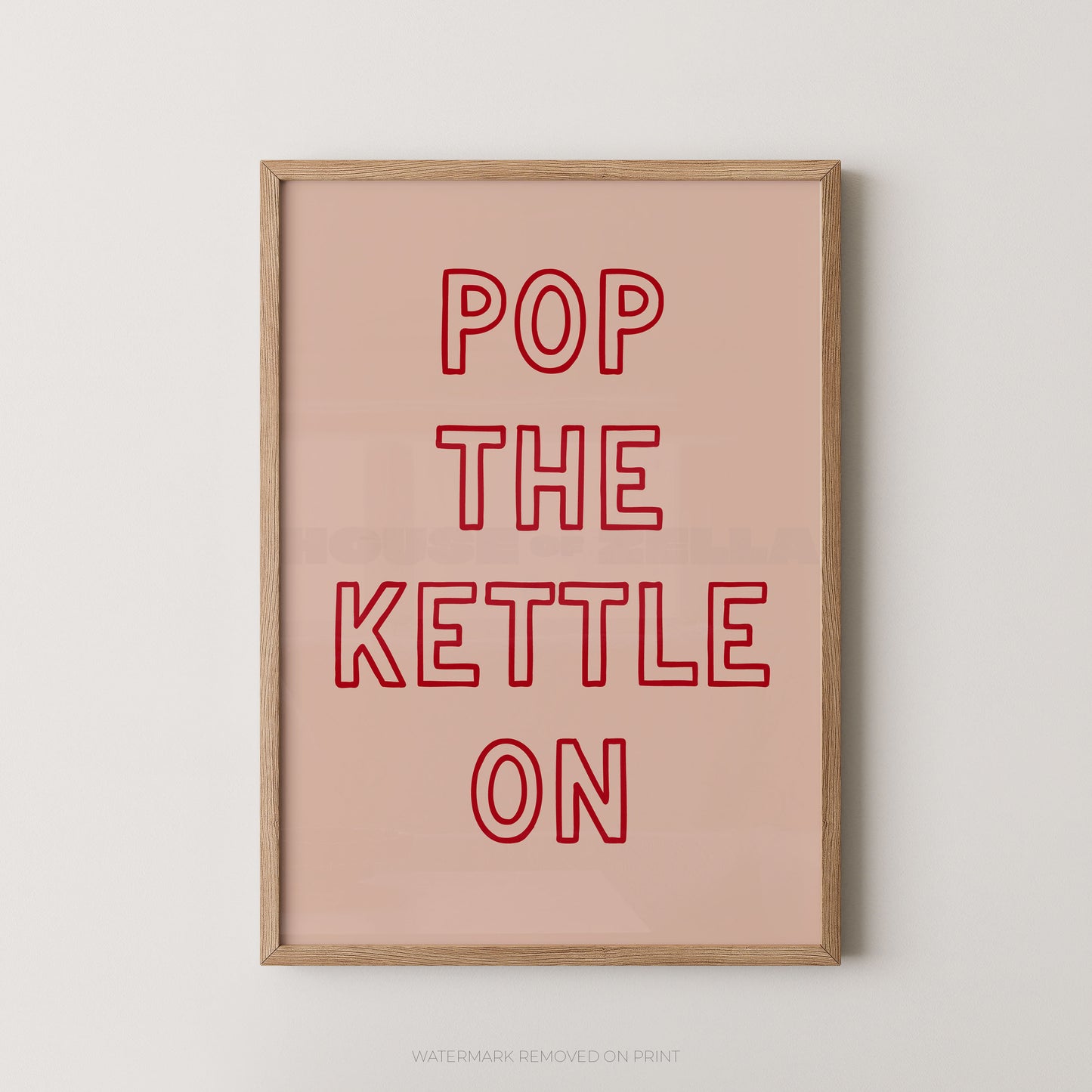 A framed typographic wall art print with the phrase “POP THE KETTLE ON” in red outlined letters on a pale pink background, shown hanging against a light neutral wall.