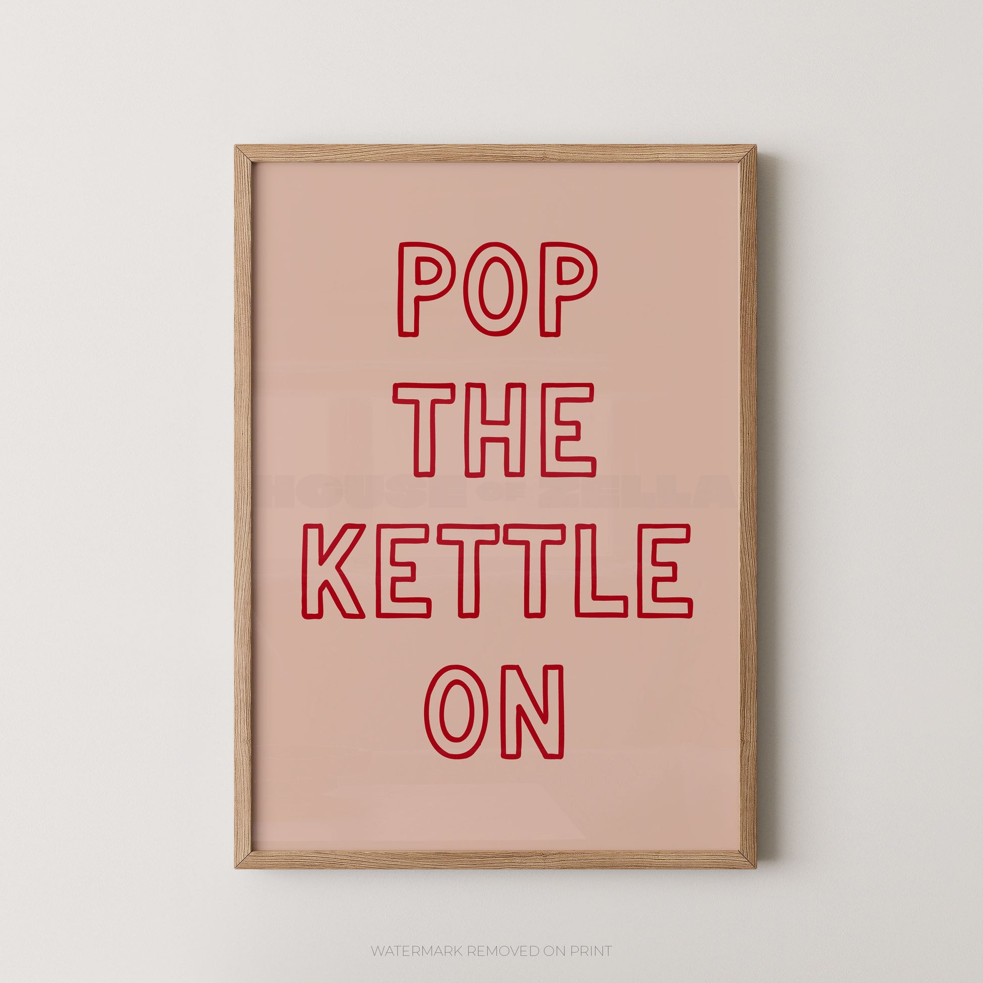 A framed typographic wall art print with the phrase “POP THE KETTLE ON” in red outlined letters on a pale pink background, shown hanging against a light neutral wall.