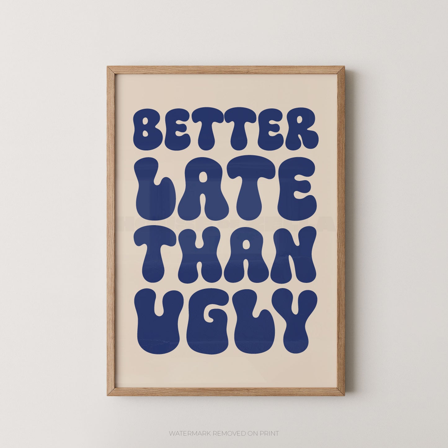 A framed typographic wall art print with the phrase “BETTER LATE THAN UGLY” in large dark blue retro letters on a warm cream background, set in a light natural wood frame against a light gray wall.