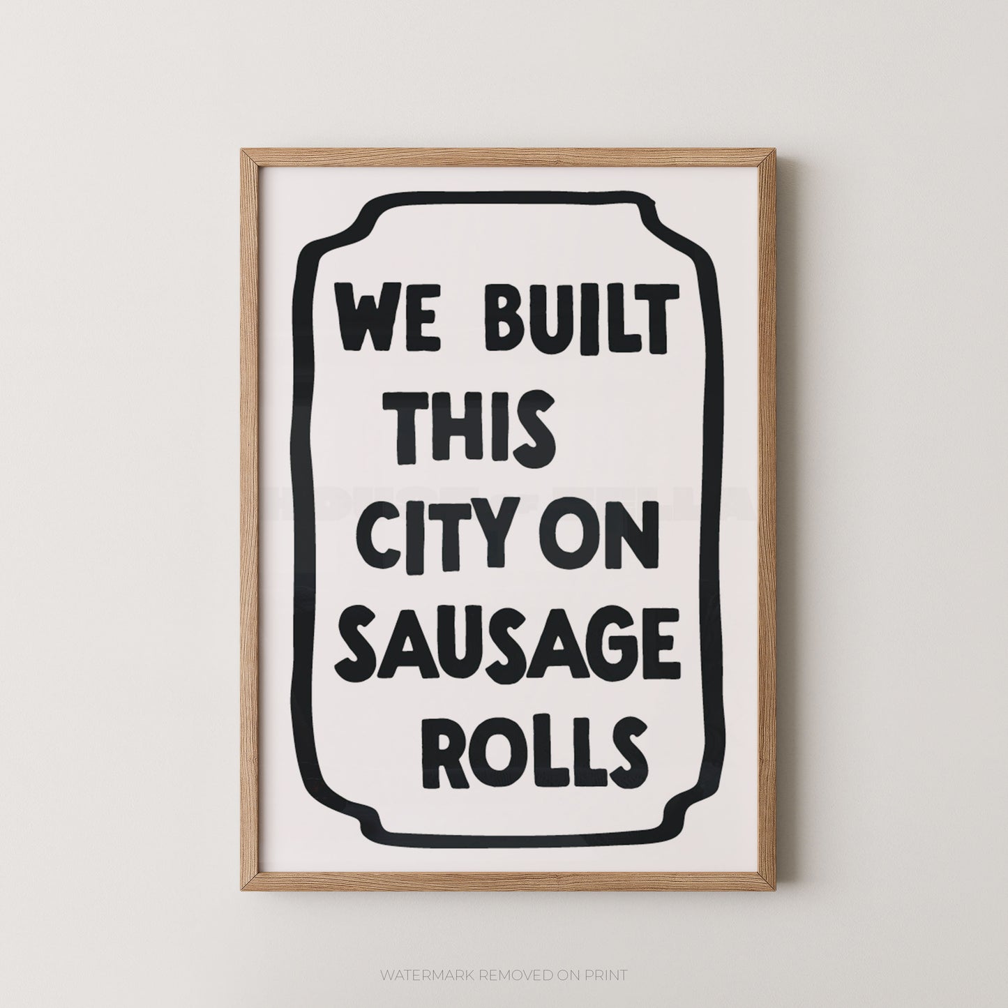 Framed wall art print with the phrase “WE BUILT THIS CITY ON SAUSAGE ROLLS” in bold black text inside a rounded black outline on a white background, shown in a light wood frame against a light gray wall.