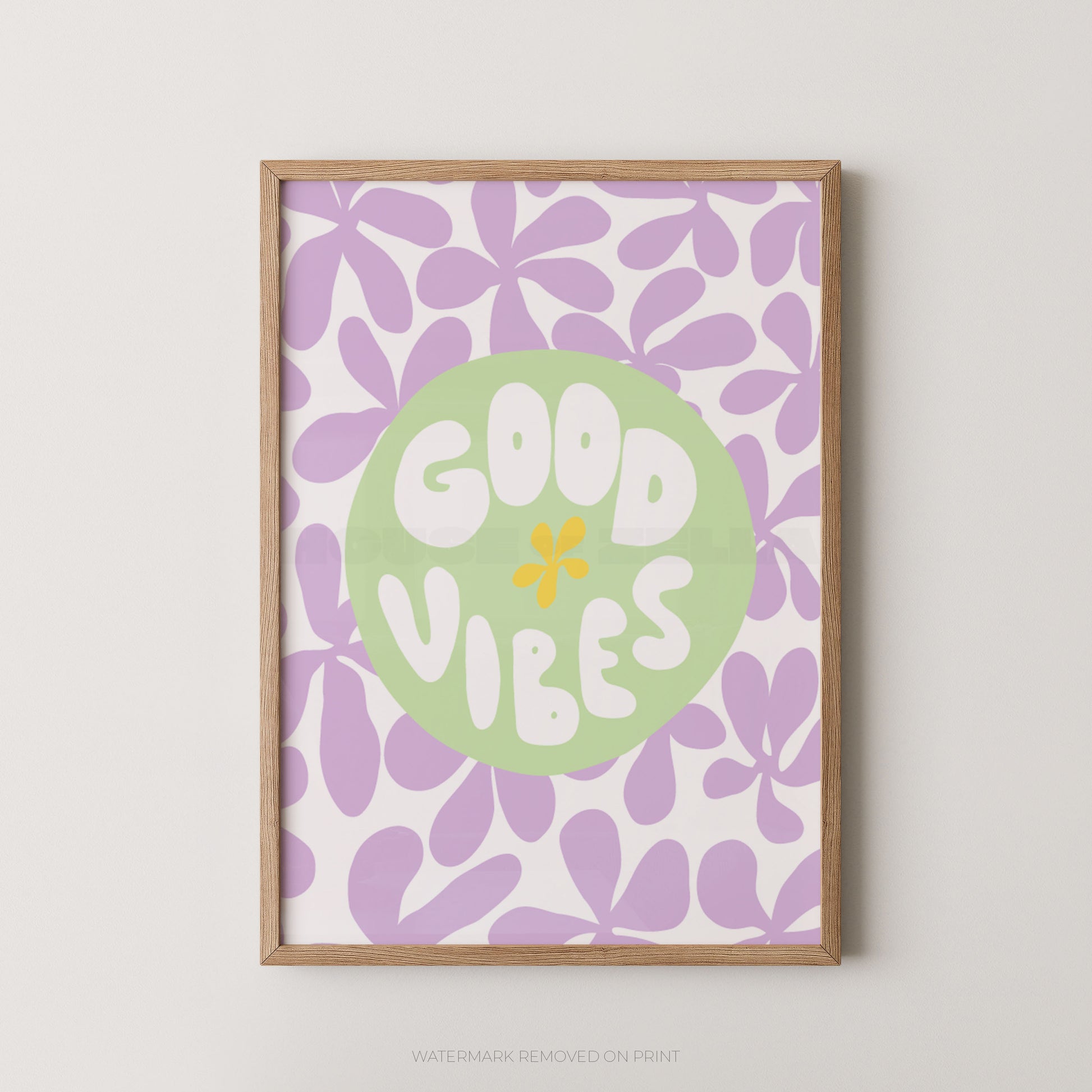 A framed art print featuring a retro floral pattern in lavender and white, with a central light green circle containing the words 'GOOD VIBES' in white letters and a small yellow flower accent. The print is housed in a light natural wood frame and is displayed against a neutral light gray wall.