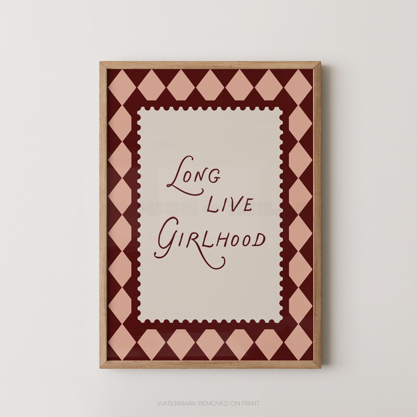 A framed typographic wall art print with the phrase “Long Live Girlhood” in dark burgundy script on a cream center panel, surrounded by a pink and burgundy diamond pattern and a light wood frame, shown against a light neutral wall.