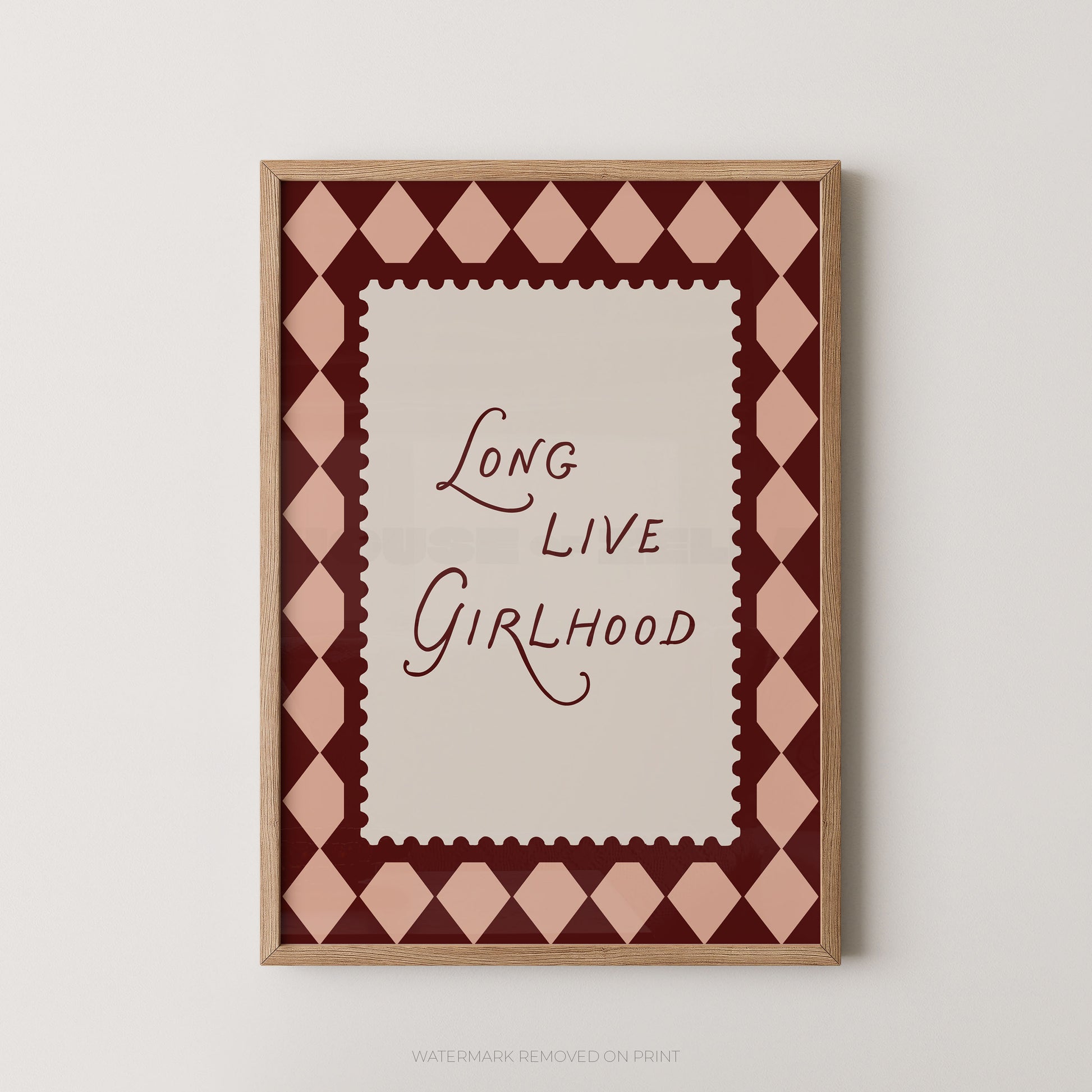 A framed typographic wall art print with the phrase “Long Live Girlhood” in dark burgundy script on a cream center panel, surrounded by a pink and burgundy diamond pattern and a light wood frame, shown against a light neutral wall.