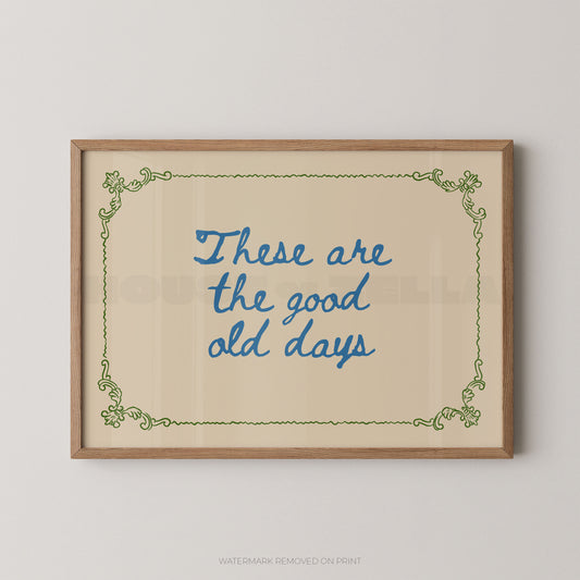 Framed wall art print with the phrase “These are the good old days” in blue script inside a light wood frame, shown against a plain light gray wall.