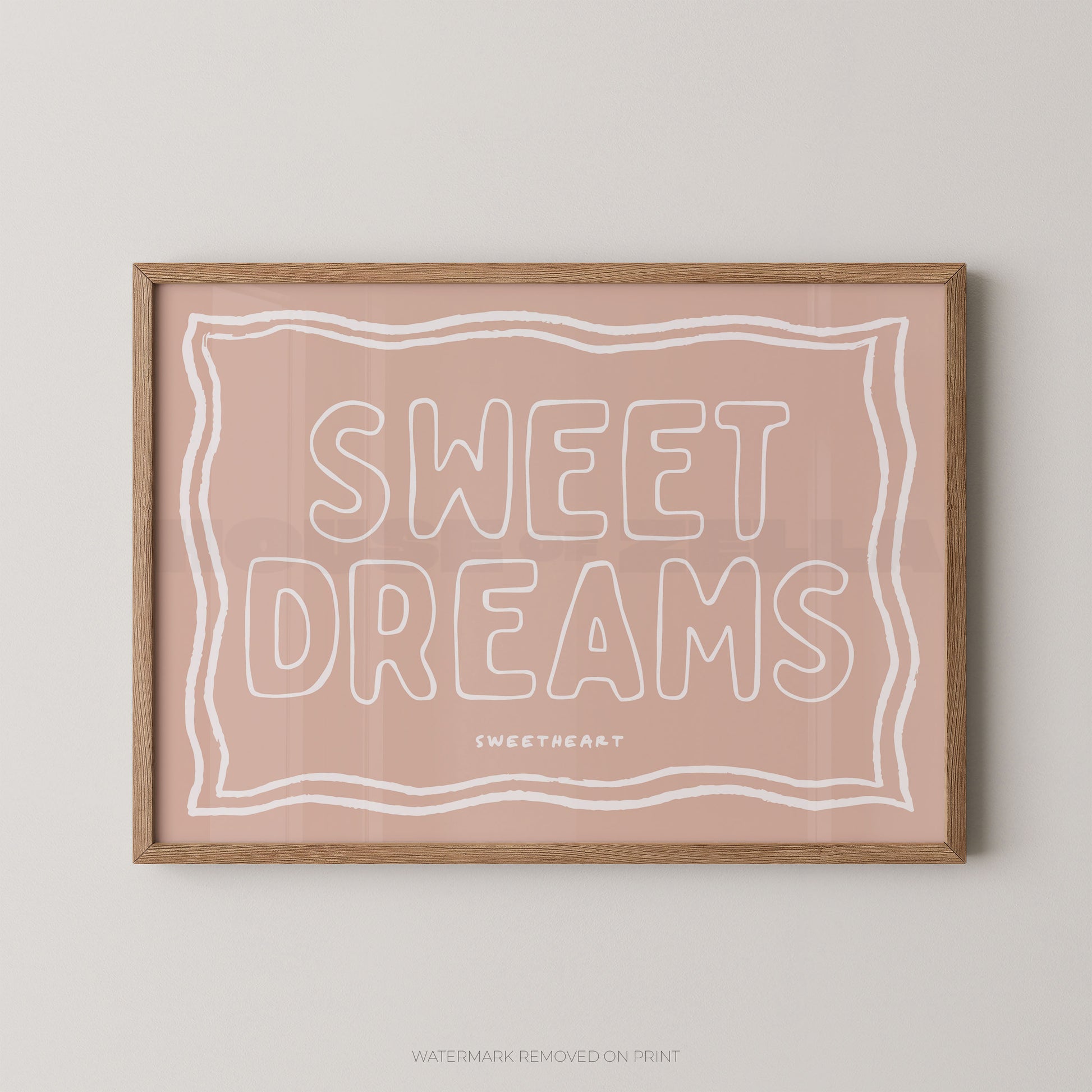 A framed art print with the words 'SWEET DREAMS' in white outline lettering on a dusty pink background, enclosed in a light natural wood frame and mounted on a light gray wall.