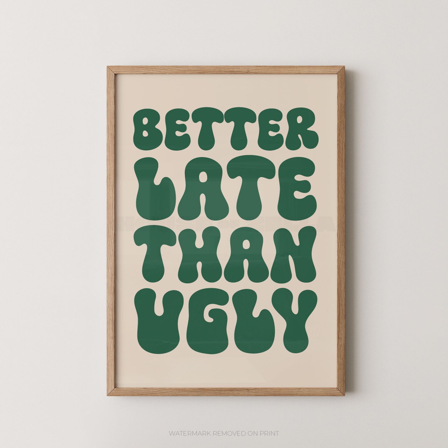 A framed typographic wall art print with the phrase “BETTER LATE THAN UGLY” in large dark green retro letters on a warm cream background, shown in a light natural wood frame against a light gray wall.