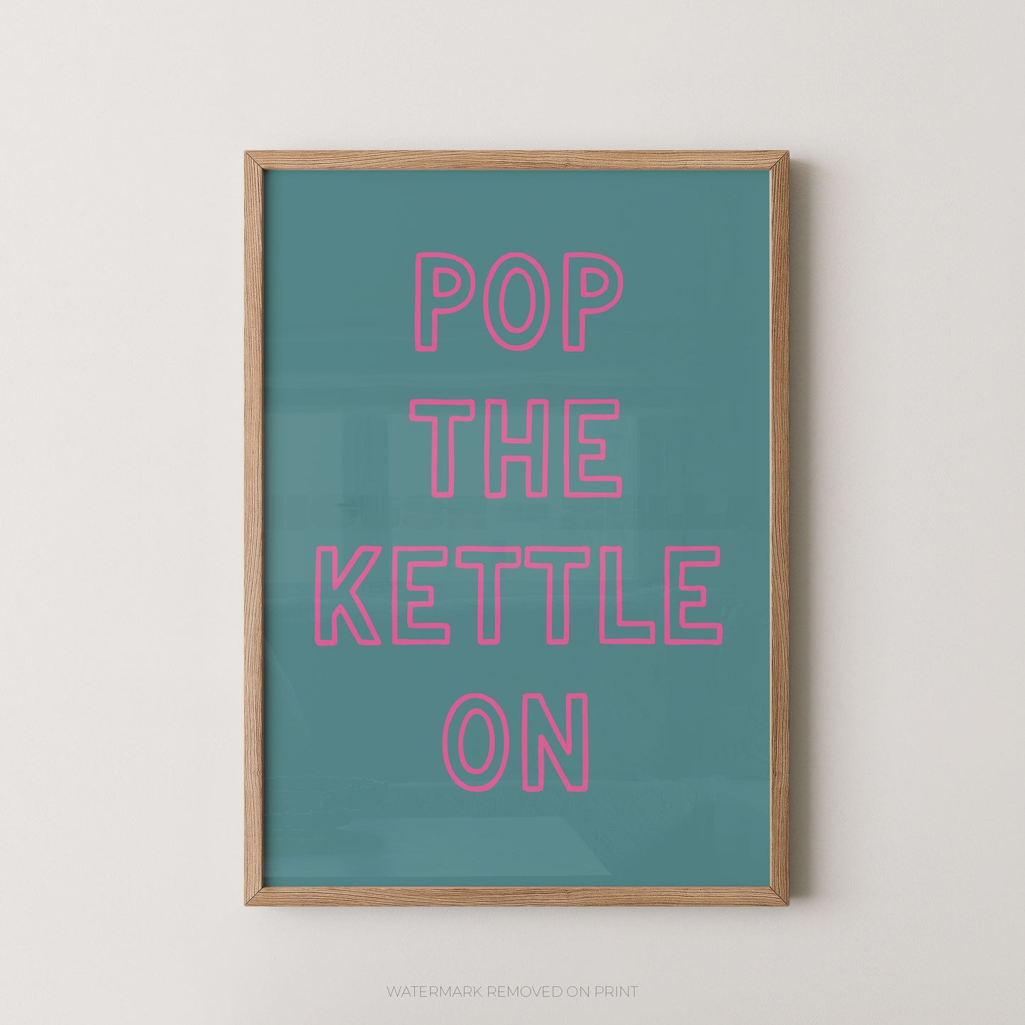 A framed typographic wall art print with the phrase “POP THE KETTLE ON” in pink outlined letters on a teal background, shown hanging against a light neutral wall.