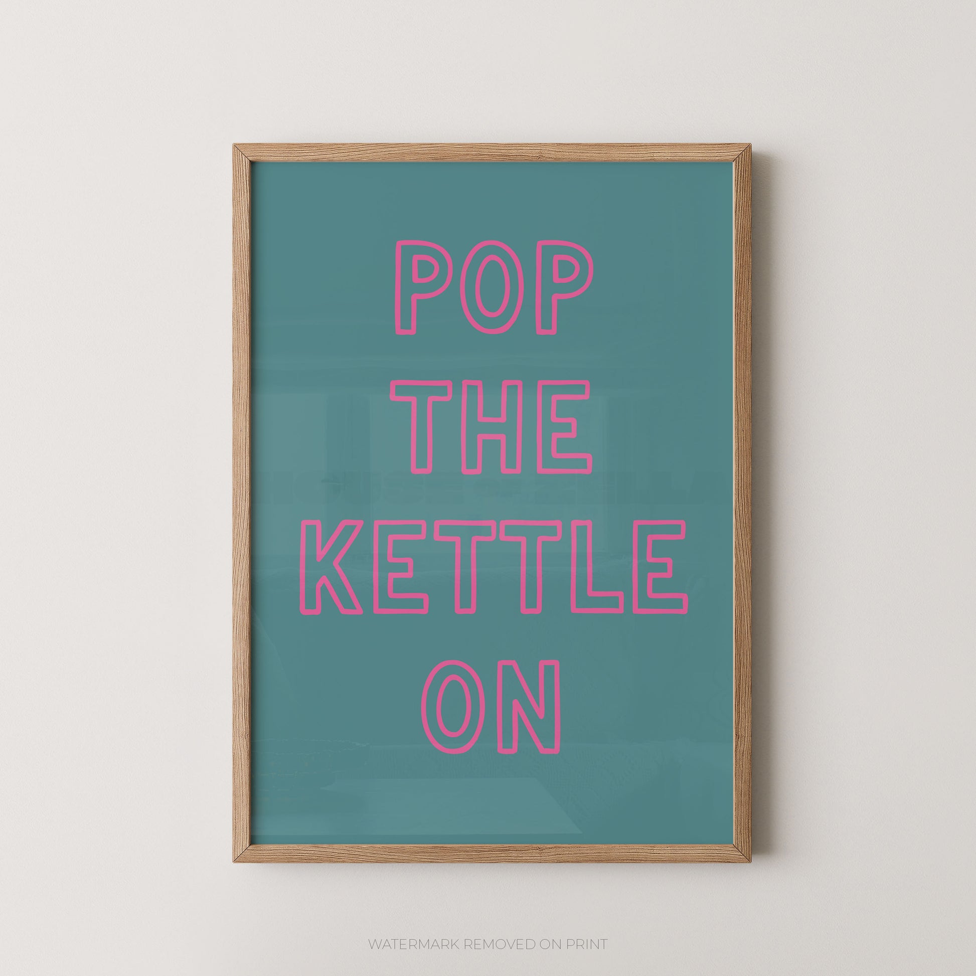 A framed typographic wall art print with the phrase “POP THE KETTLE ON” in pink outlined letters on a teal background, shown hanging against a light neutral wall.