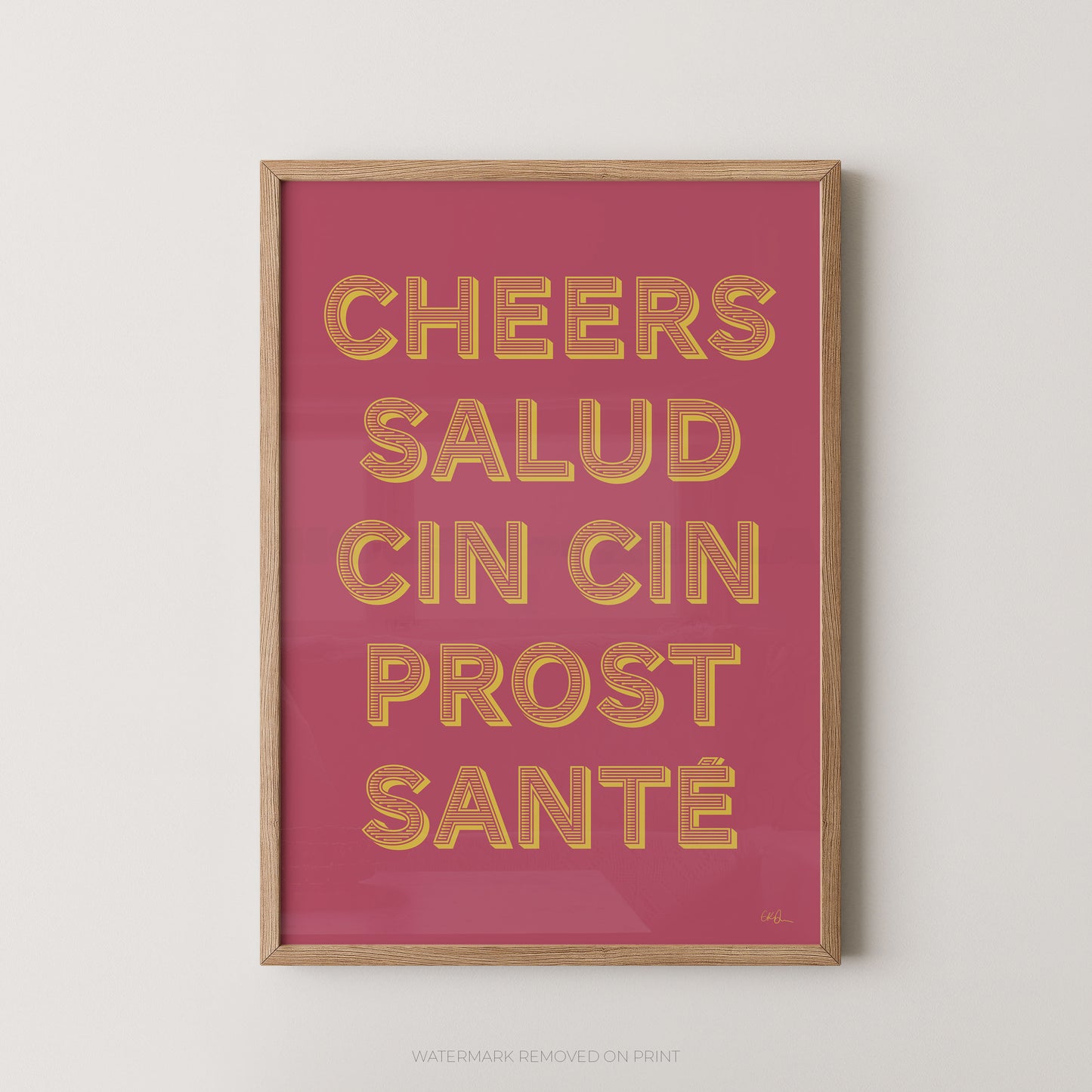 Framed wall art print with the phrase “CHEERS SALUD CIN CIN PROST SANTÉ” in raised gold letters on a pink background, shown in a light wood frame against a light gray wall.