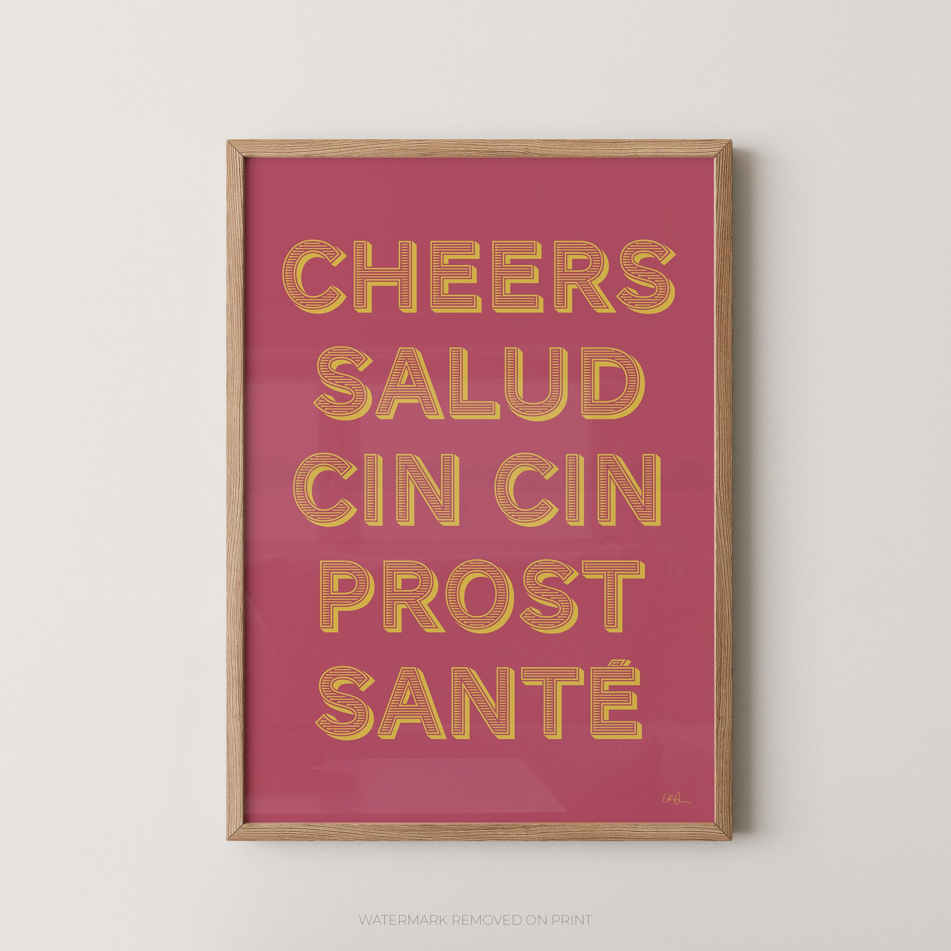 Framed wall art print with the phrase “CHEERS SALUD CIN CIN PROST SANTÉ” in raised gold letters on a pink background, shown in a light wood frame against a light gray wall.