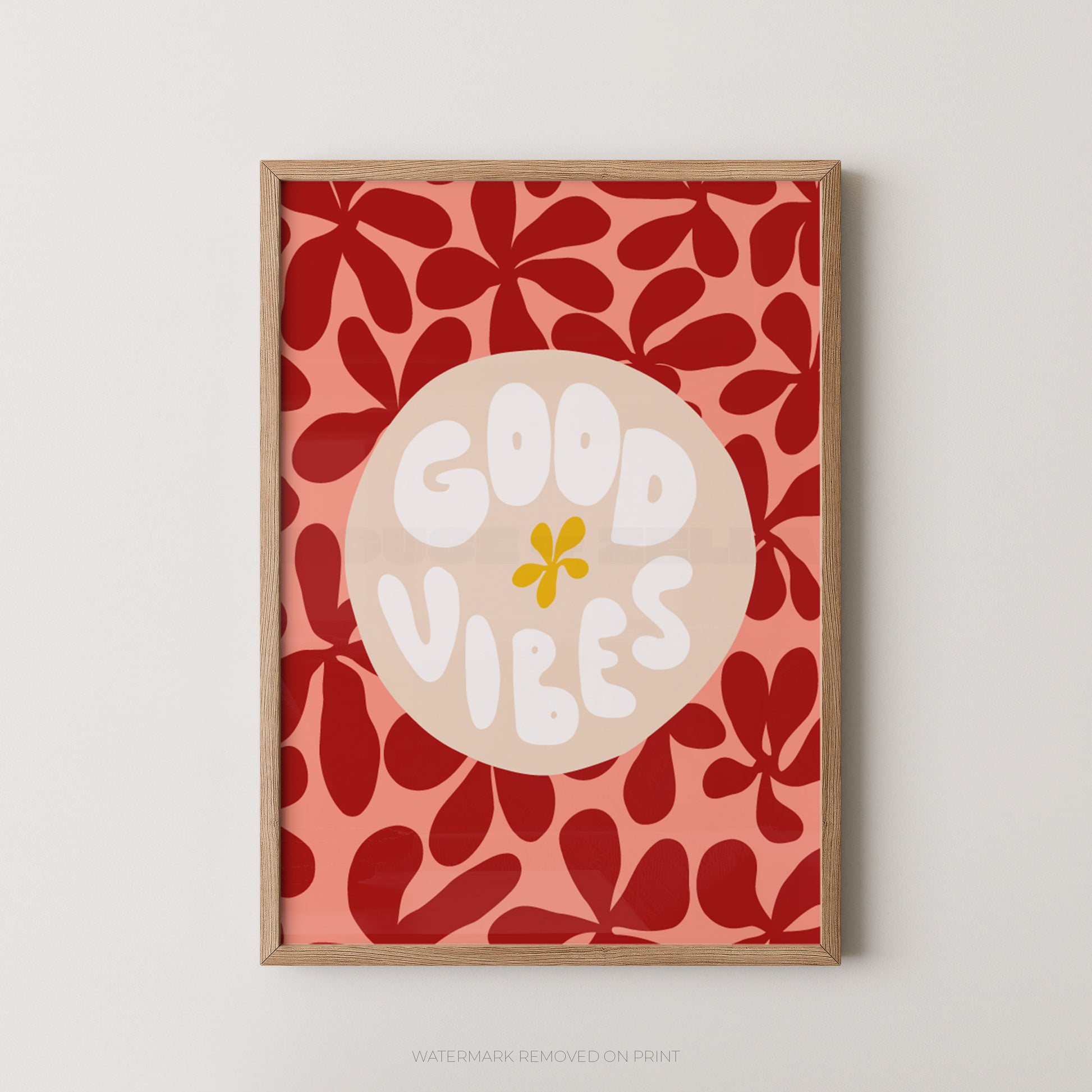 A framed art print featuring a retro floral pattern in red and coral pink, with a central beige circle containing the words 'GOOD VIBES' in white and a small yellow flower accent. The print is housed in a light natural wood frame and is displayed against a neutral light gray wall.