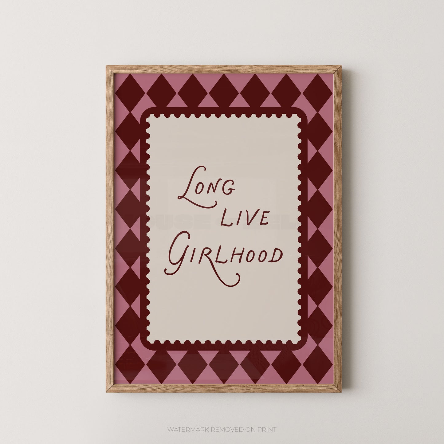 A framed art print featuring the text 'Long Live Girlhood' in dark red script on a cream background, surrounded by a pink and dark red diamond pattern, in a light wood frame.