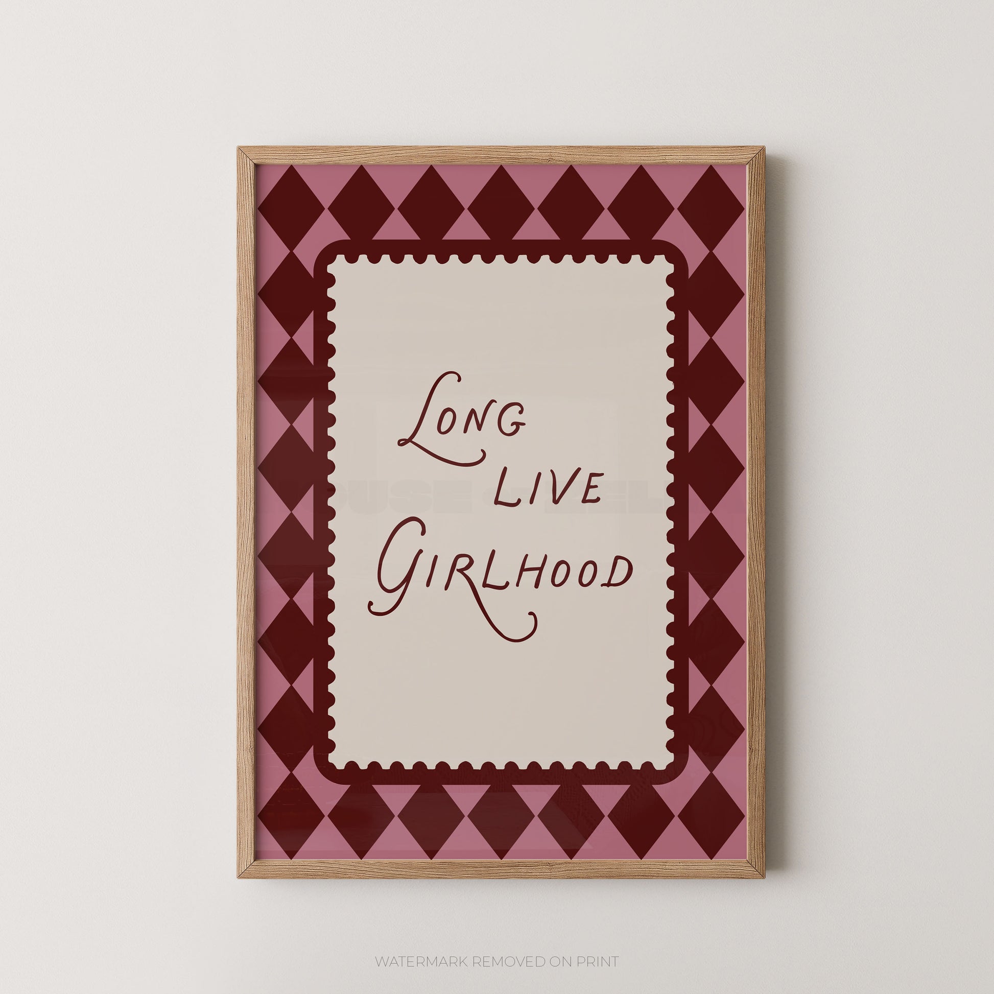 A framed art print featuring the text 'Long Live Girlhood' in dark red script on a cream background, surrounded by a pink and dark red diamond pattern, in a light wood frame.