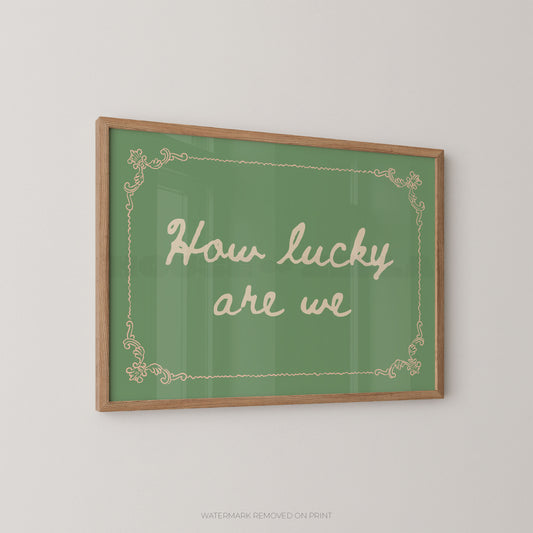 A framed wall art print with a green background, cream script text reading 'How lucky are we,' and a light wood frame, shown hanging on a plain light wall.