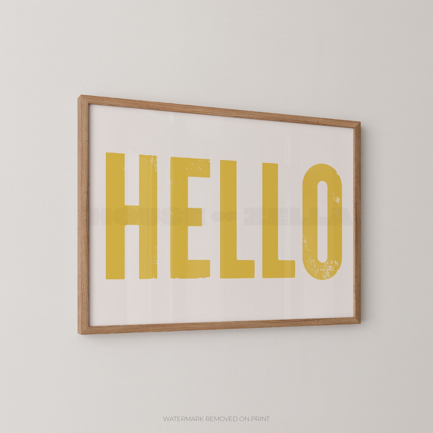Framed wall art print with the word "HELLO" in large mustard-yellow letters on a white background, set in a light natural wood frame against a light gray wall.