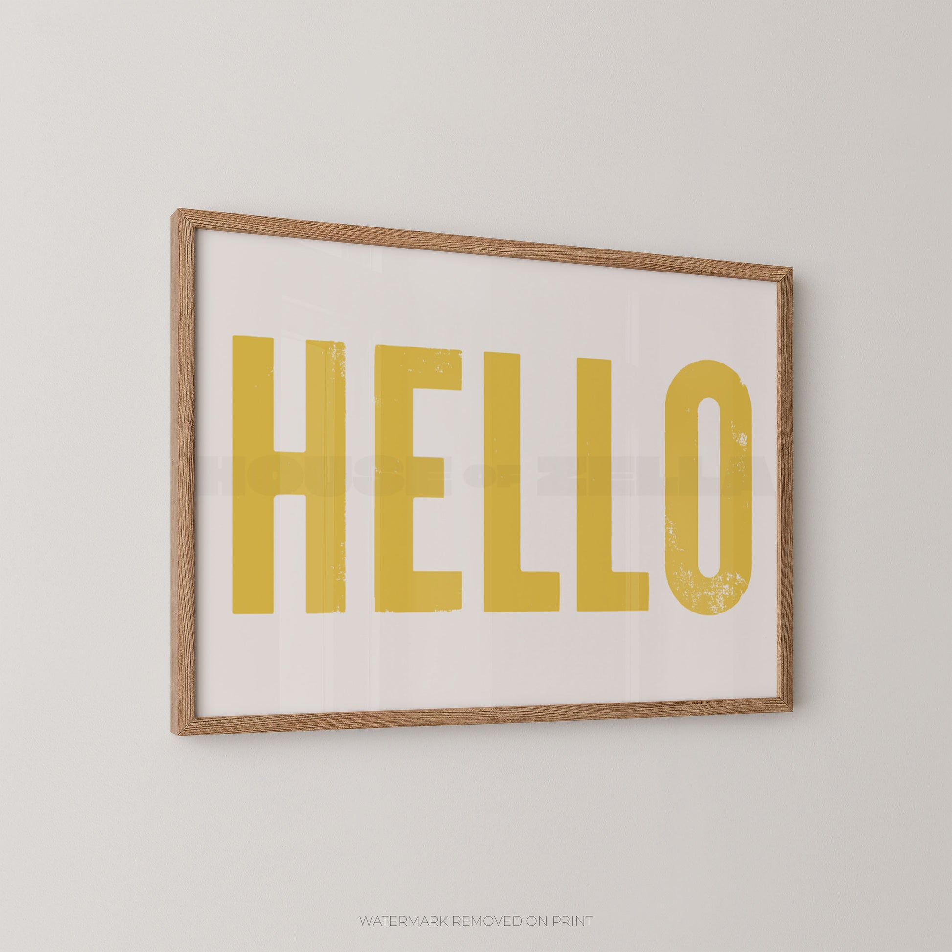 Framed wall art print with the word "HELLO" in large mustard-yellow letters on a white background, set in a light natural wood frame against a light gray wall.