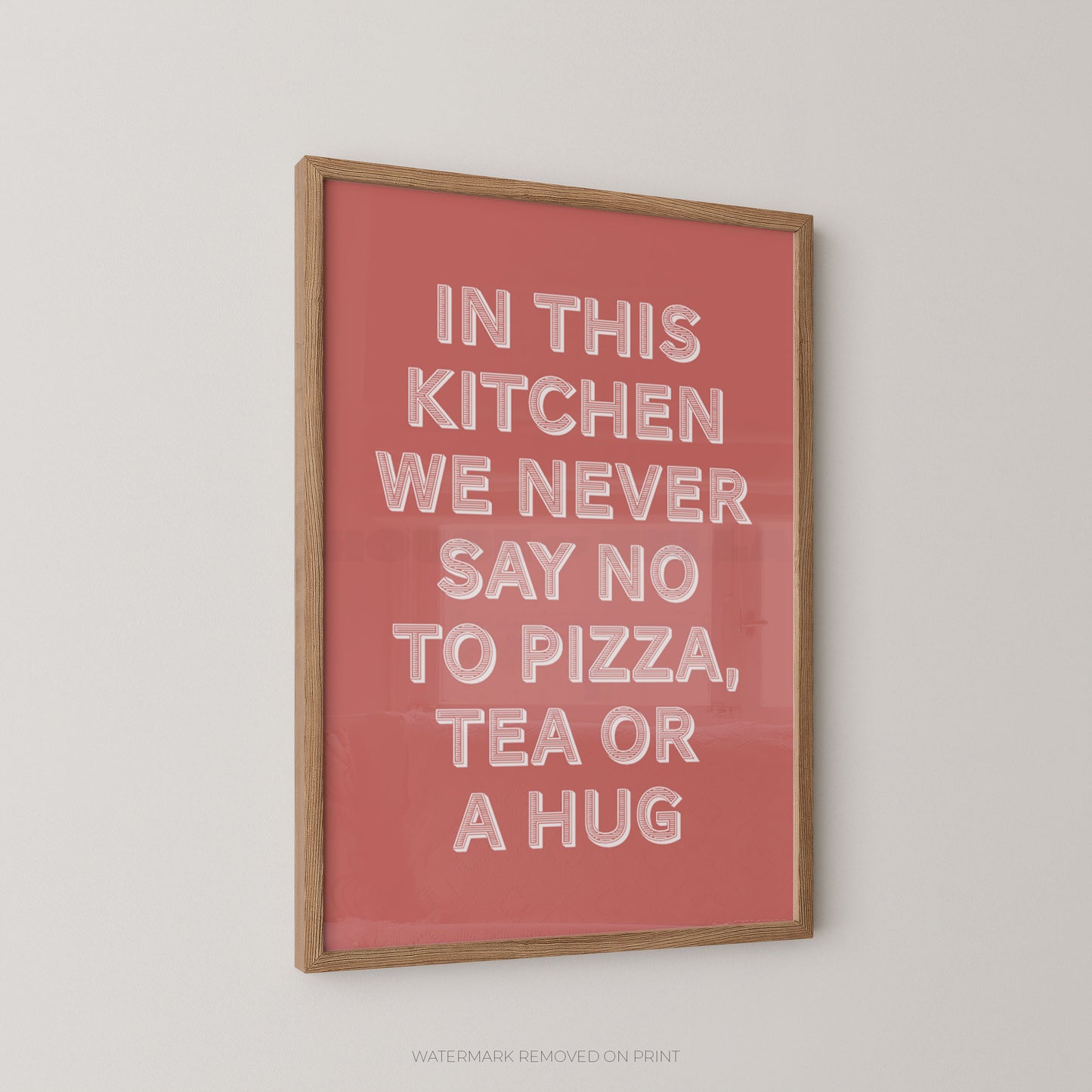 A framed typographic wall art print with a coral pink background and white raised lettering that reads, 'IN THIS KITCHEN WE NEVER SAY NO TO PIZZA, TEA OR A HUG,' shown hanging on a light neutral wall.