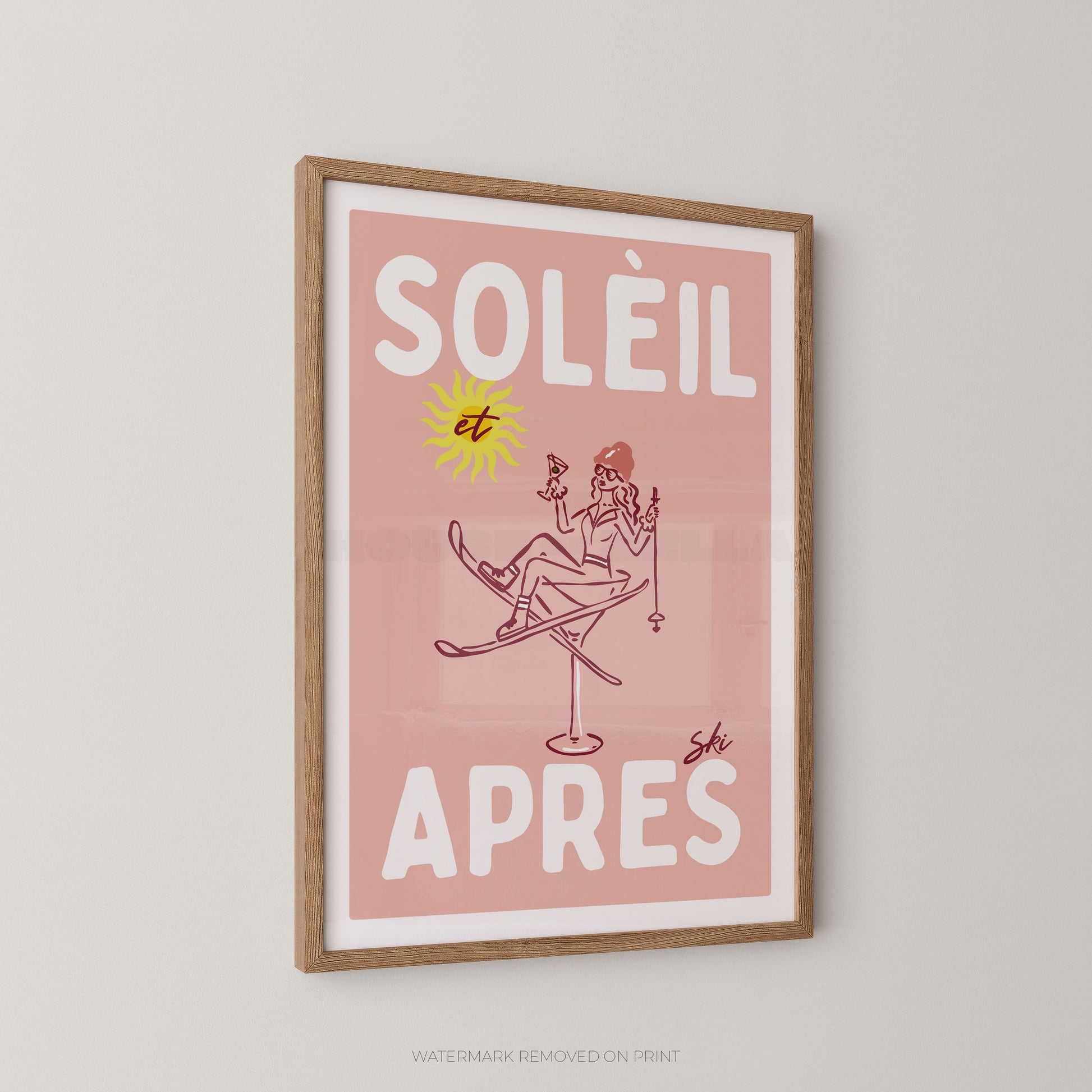 A framed art print featuring a retro illustration with the French text 'SOLÉIL et APRES' on a dusty pink background. The print includes a yellow sun and a line-drawn figure sitting on skis, all enclosed in a light wood frame and displayed on a neutral light gray wall.