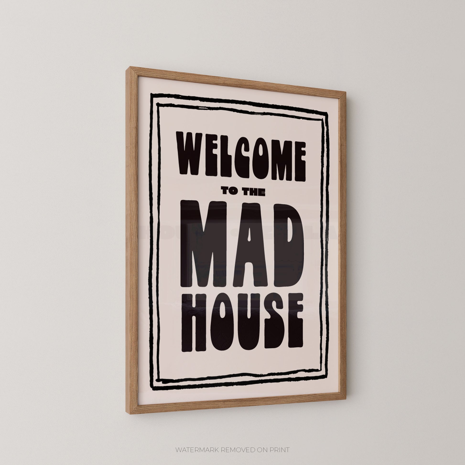 Framed wall art print with the text 'WELCOME TO THE MAD HOUSE' in bold black lettering on a cream background, enclosed in a light wood frame and shown hanging on a light gray wall.