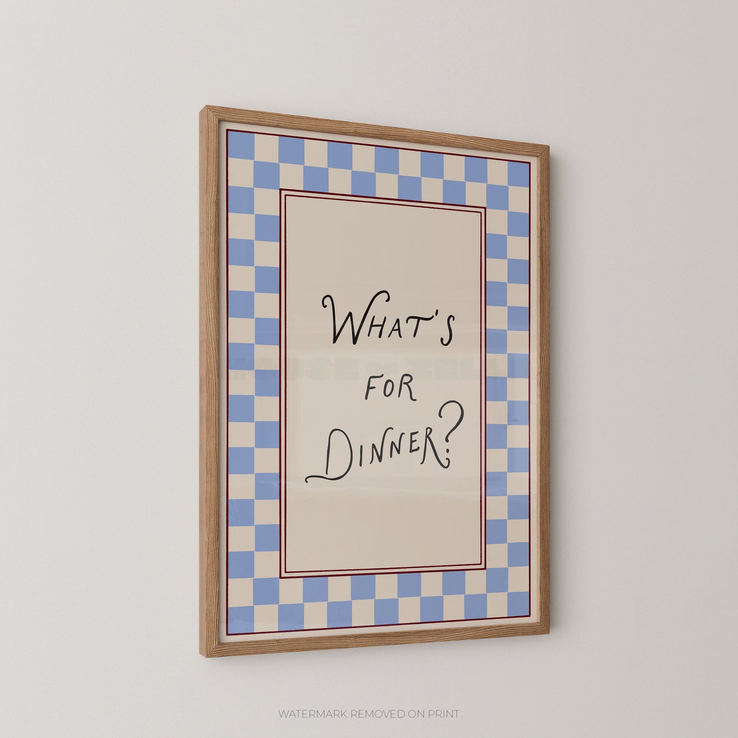 A framed typographic wall art print hanging on a light gray wall. The print has a blue-and-white checkered border and a cream center panel with the handwritten text “WHAT’S FOR DINNER?” in black script.