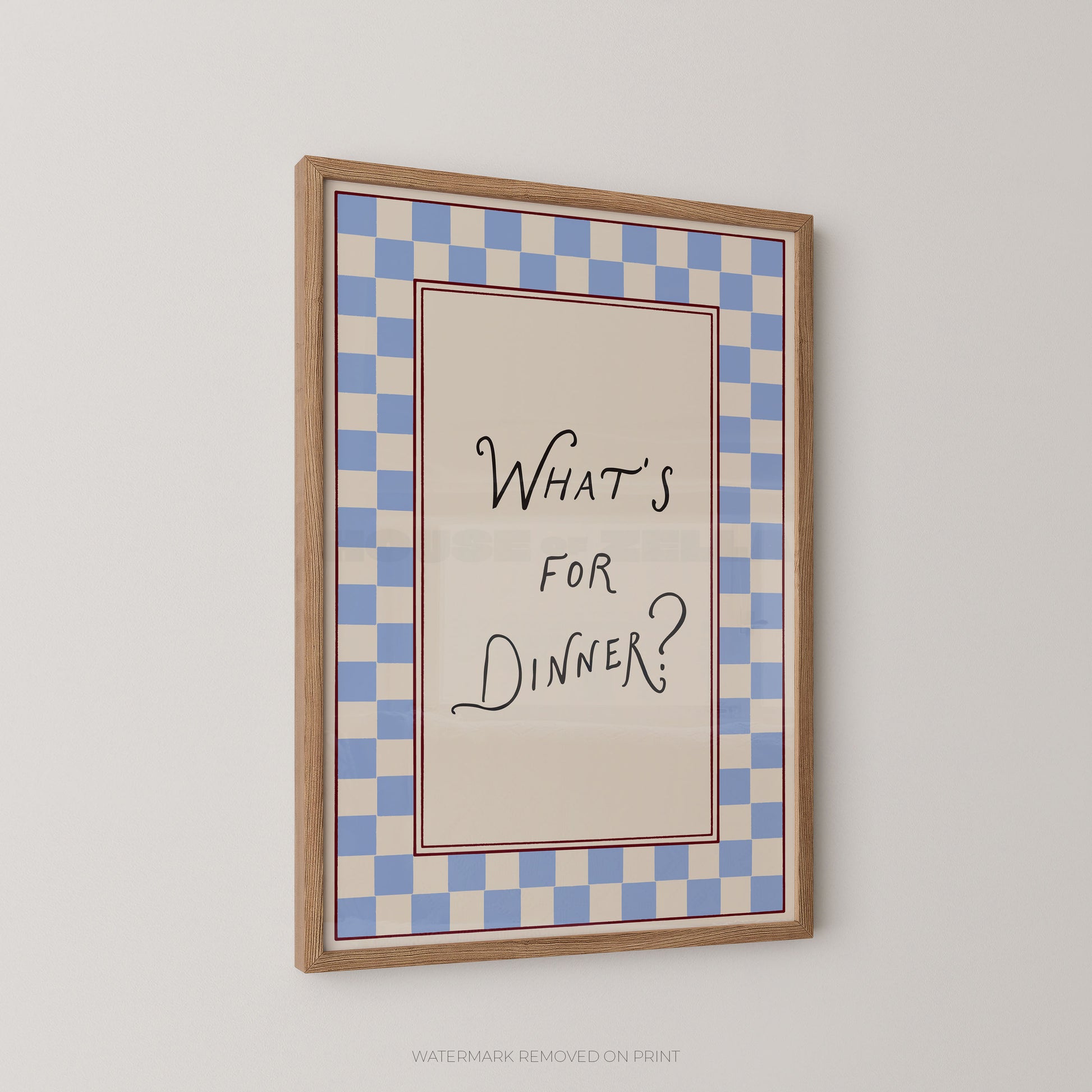 A framed typographic wall art print hanging on a light gray wall. The print has a blue-and-white checkered border and a cream center panel with the handwritten text “WHAT’S FOR DINNER?” in black script.