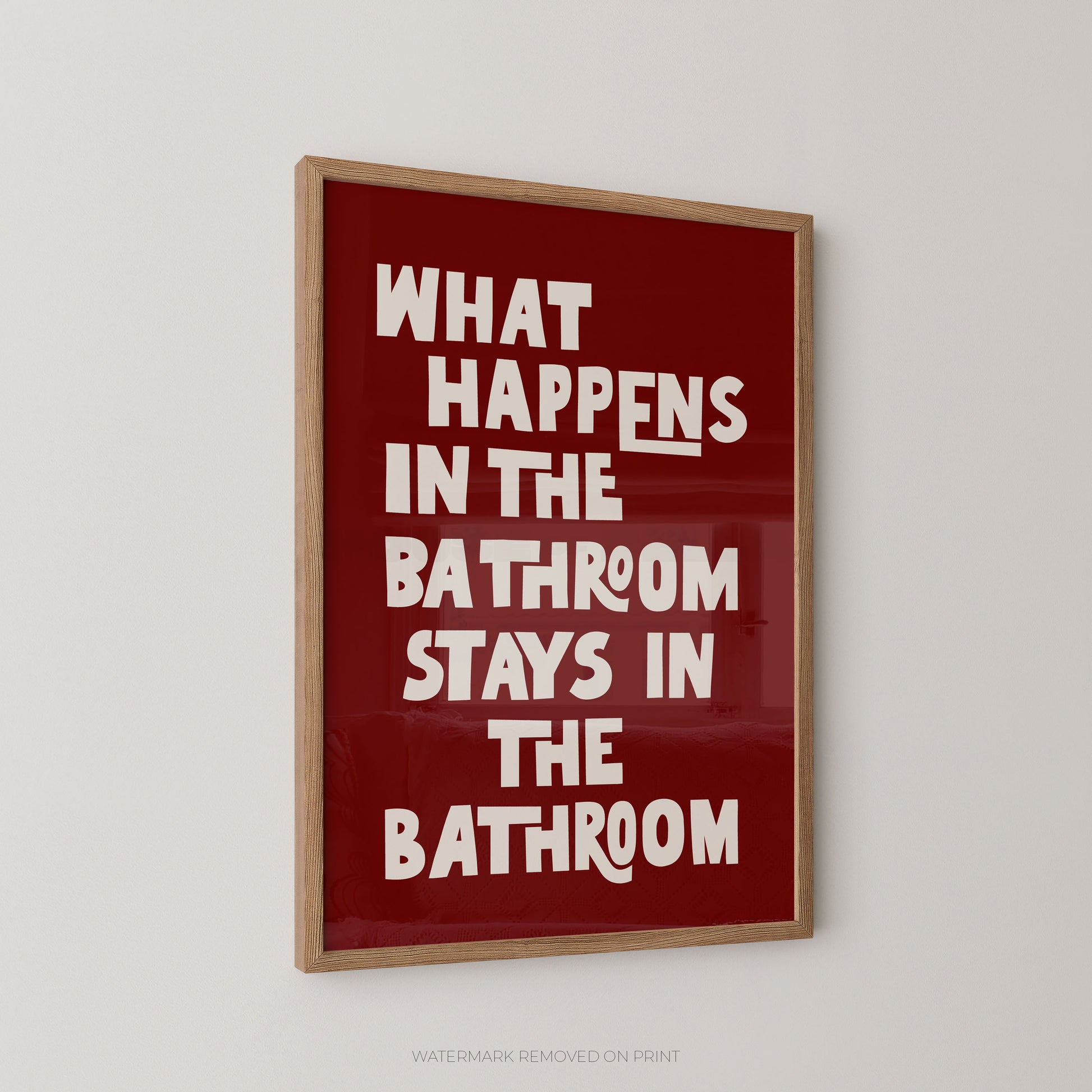 Funny bathroom wall art print in a wood frame – playful chunky cream typography on a dark red background reading "What Happens In The Bathroom Stays In The Bathroom"