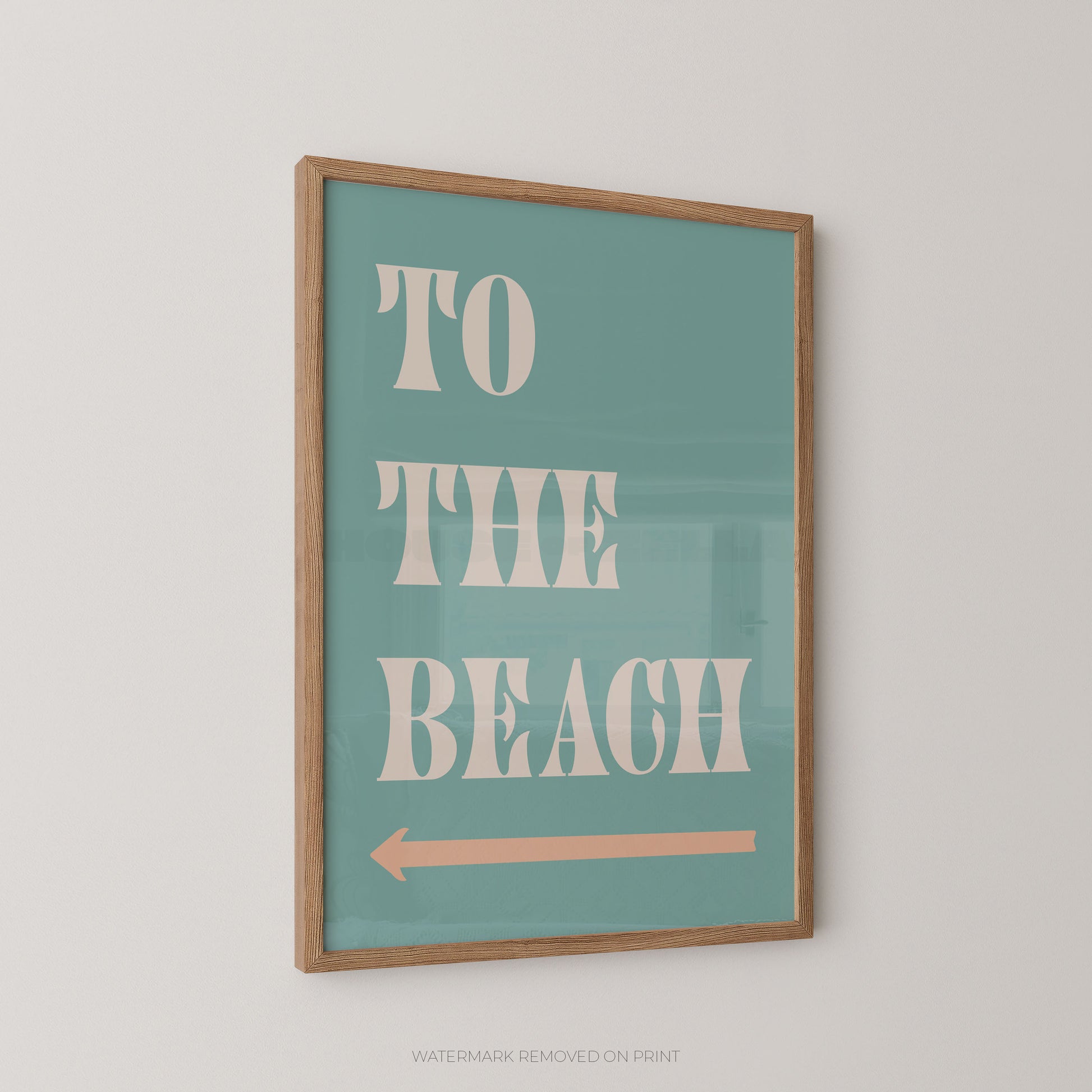 A framed typographic wall art print with the words “TO THE BEACH” in large cream letters on a muted teal background, with a peach arrow beneath. The print is shown hanging on a light neutral wall.
