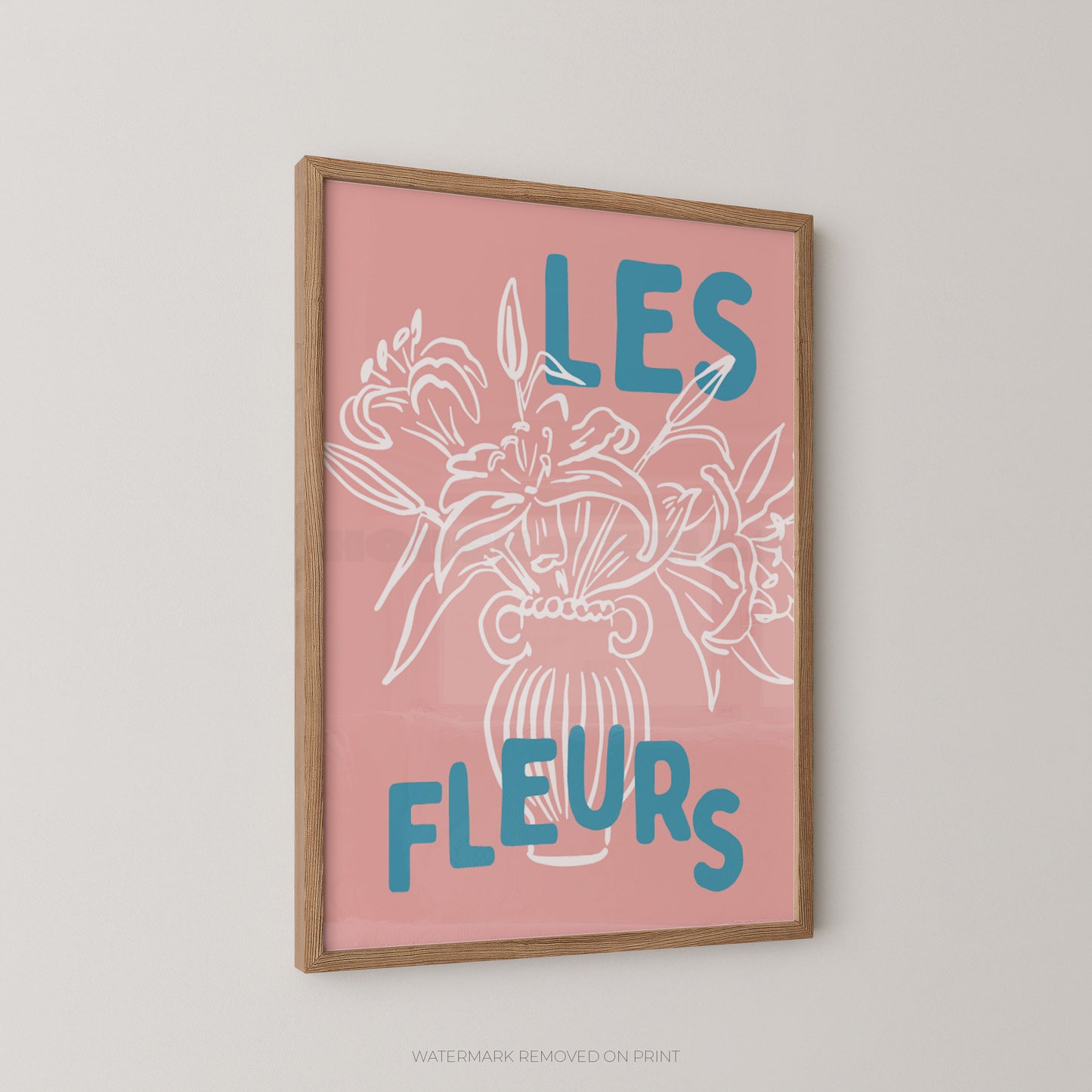 A framed wall art print with a pink background, white floral line drawing, and blue text reading “LES FLEURS,” shown hanging on a light neutral wall.
