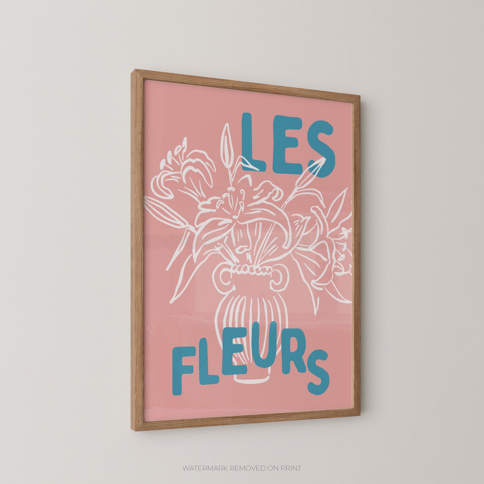 A framed wall art print with a pink background, white floral line drawing, and blue text reading “LES FLEURS,” shown hanging on a light neutral wall.