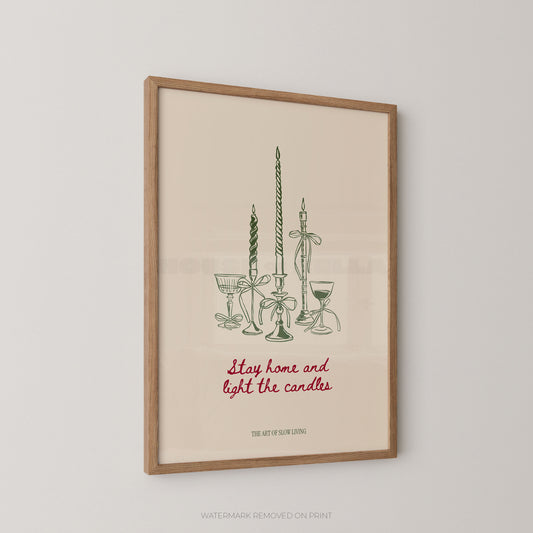 Framed art print featuring a minimalist illustration of candlesticks and glassware with the phrase 'Stay home and light the candles' in red script, shown hanging on a light gray wall.