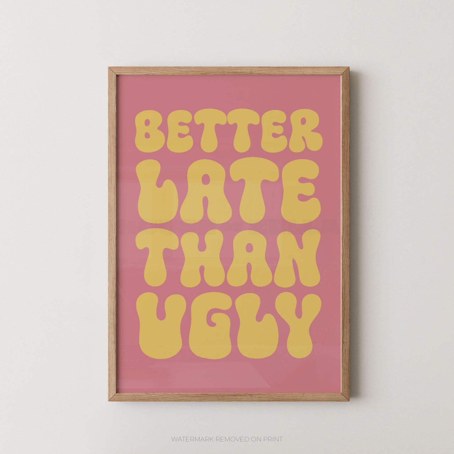 A framed typographic wall art print with the phrase “BETTER LATE THAN UGLY” in large yellow retro letters on a pink background, shown hanging against a light neutral wall.