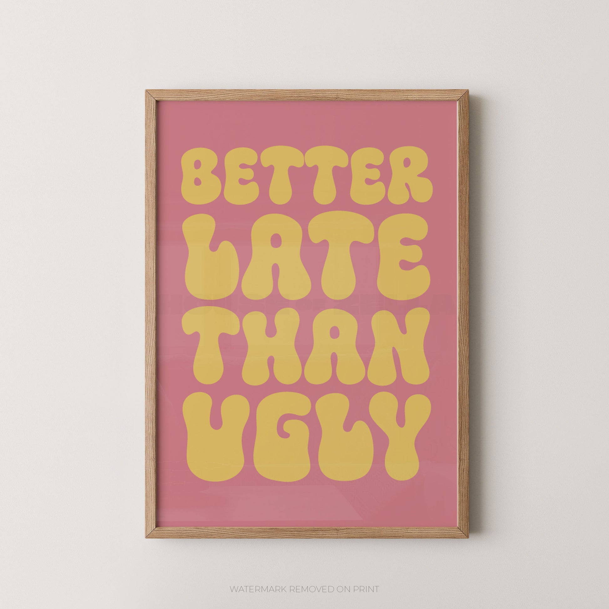 A framed typographic wall art print with the phrase “BETTER LATE THAN UGLY” in large yellow retro letters on a pink background, shown hanging against a light neutral wall.