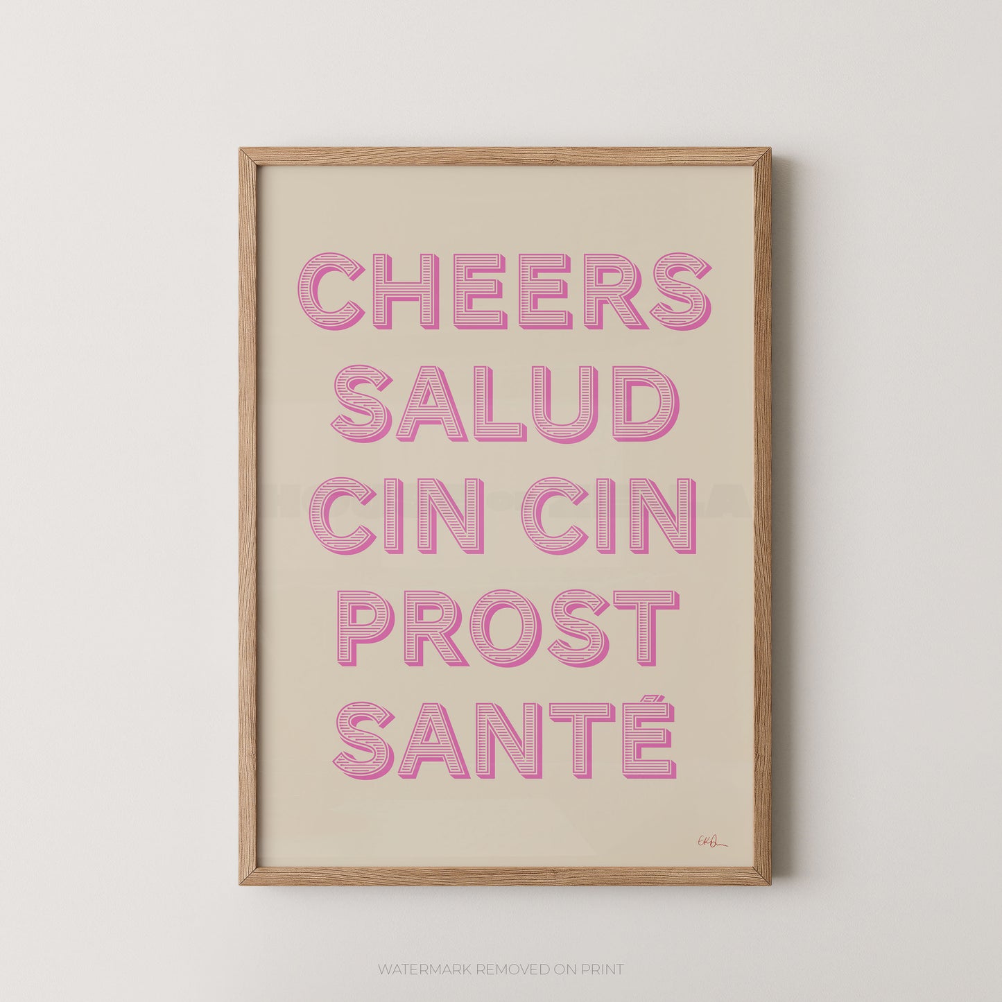A framed typographic wall art print with the phrase “CHEERS SALUD CIN CIN PROST SANTÉ” in pink raised letters on a warm cream background, shown in a light natural wood frame against a light gray wall.
