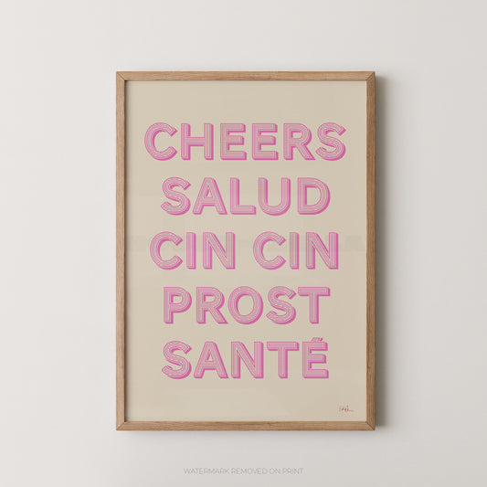 A framed typographic wall art print with the phrase “CHEERS SALUD CIN CIN PROST SANTÉ” in pink raised letters on a warm cream background, shown in a light natural wood frame against a light gray wall.