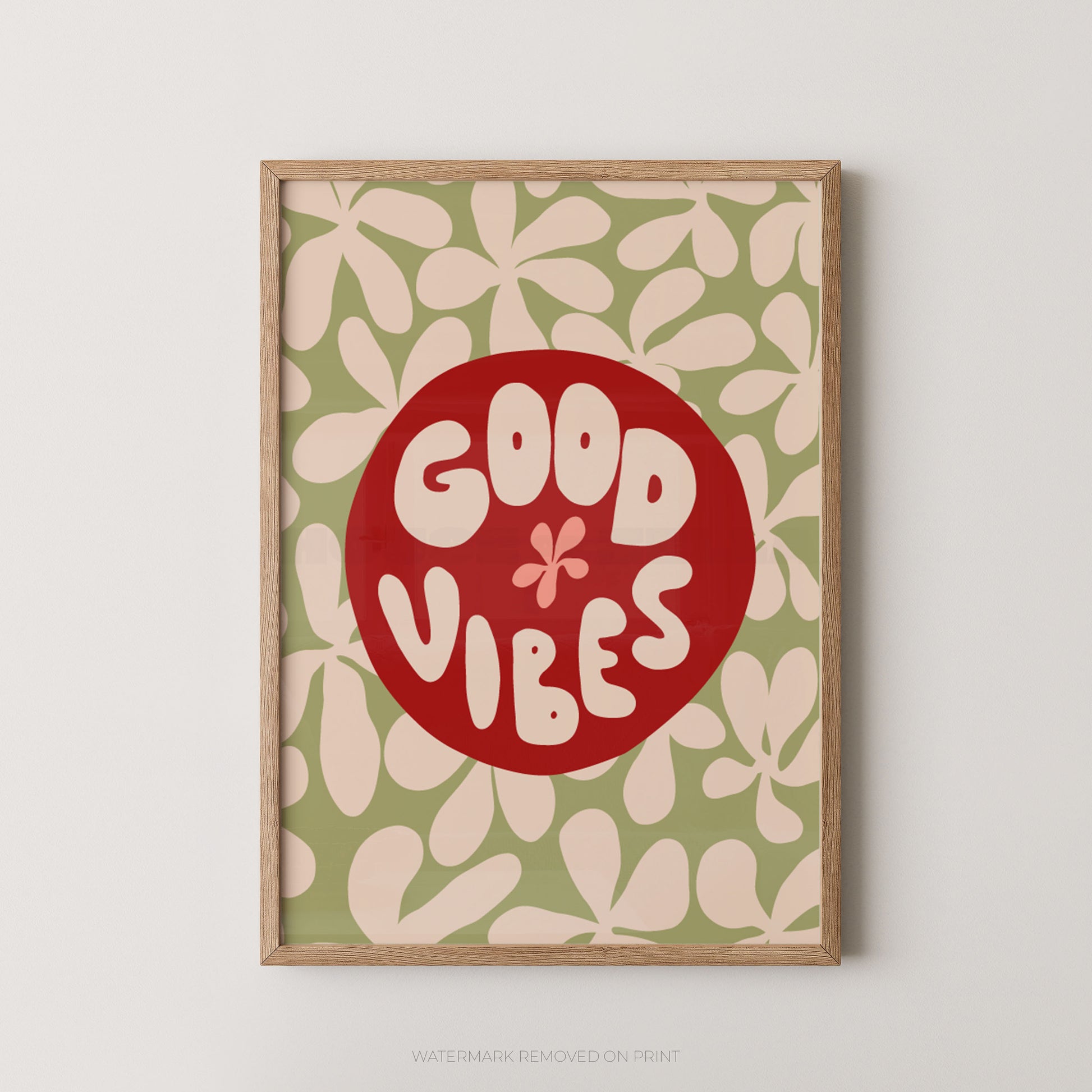 A framed art print featuring a retro design with a red circle containing the words 'GOOD VIBES' in cream lettering, set against a sage green background with a cream floral pattern. The print is housed in a light natural wood frame and is displayed on a neutral light gray wall.
