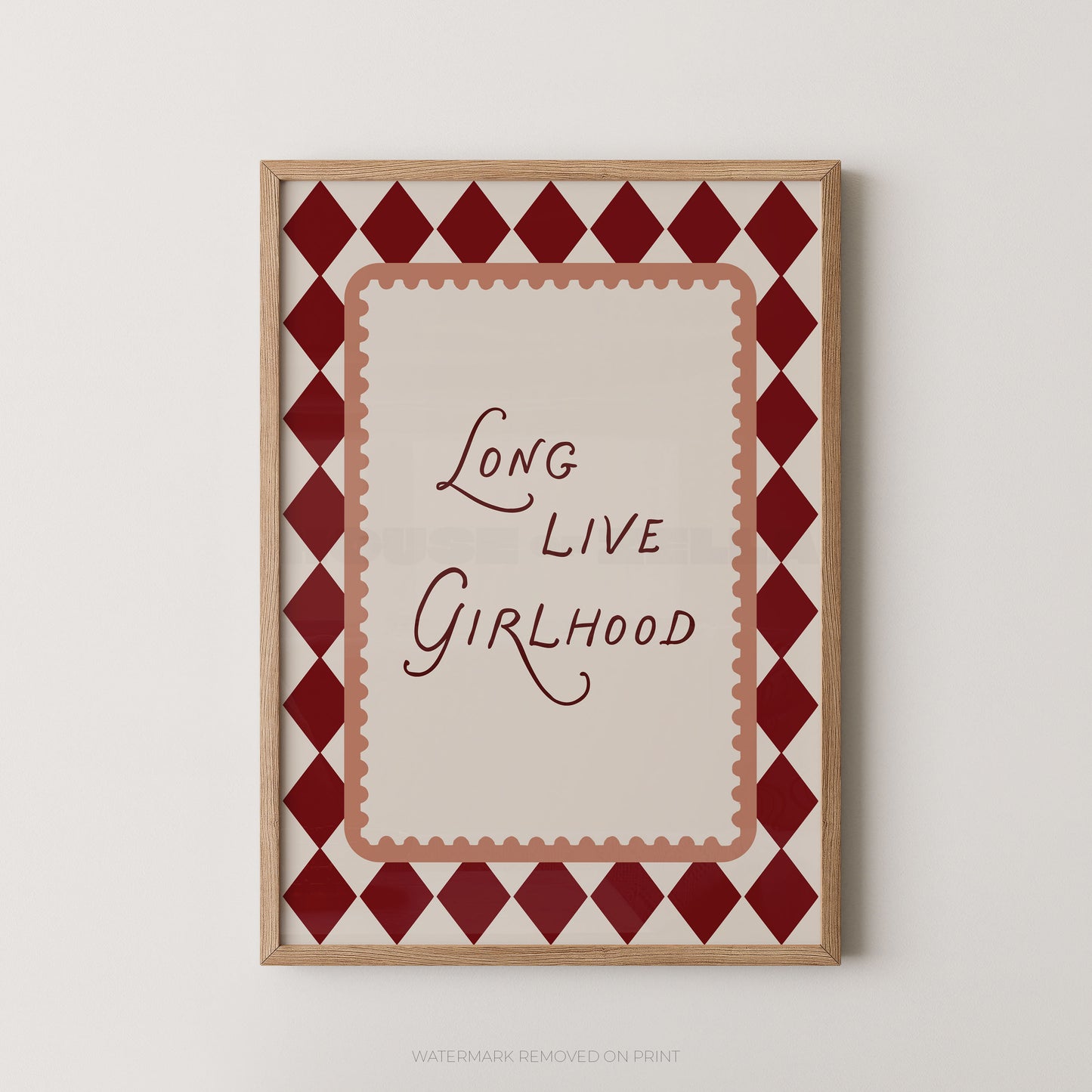 A framed art print featuring the phrase 'Long Live Girlhood' in dark red script on a cream background, surrounded by a red and cream harlequin diamond border and a light wood frame, shown against a light gray wall.