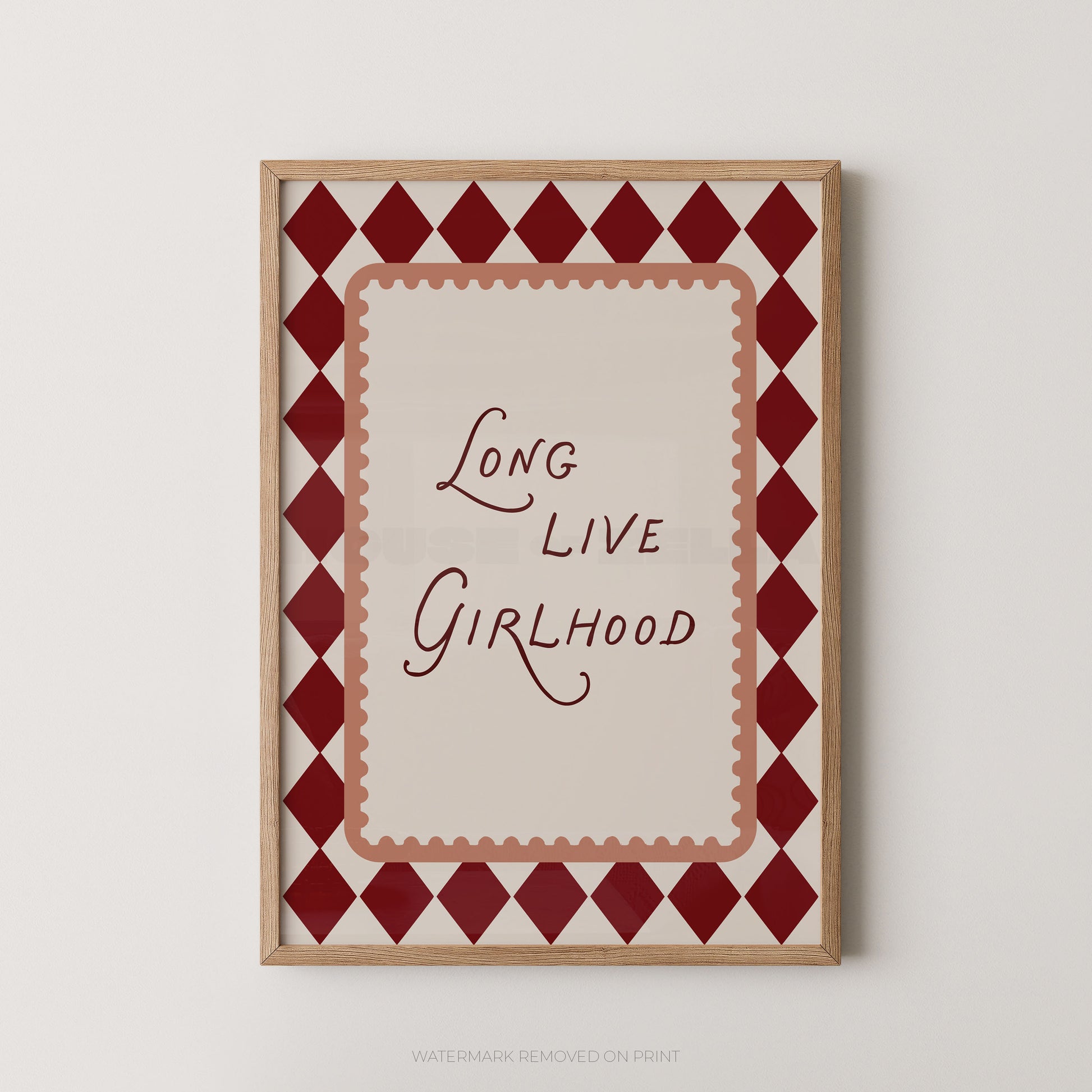 A framed art print featuring the phrase 'Long Live Girlhood' in dark red script on a cream background, surrounded by a red and cream harlequin diamond border and a light wood frame, shown against a light gray wall.
