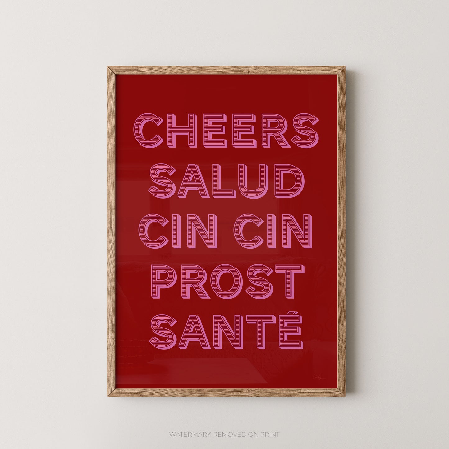 Framed wall art print with the phrase 'CHEERS SALUD CIN CIN PROST SANTÉ' in pink neon-style outlined lettering on a deep red background, shown in a light wood frame against a light gray wall.