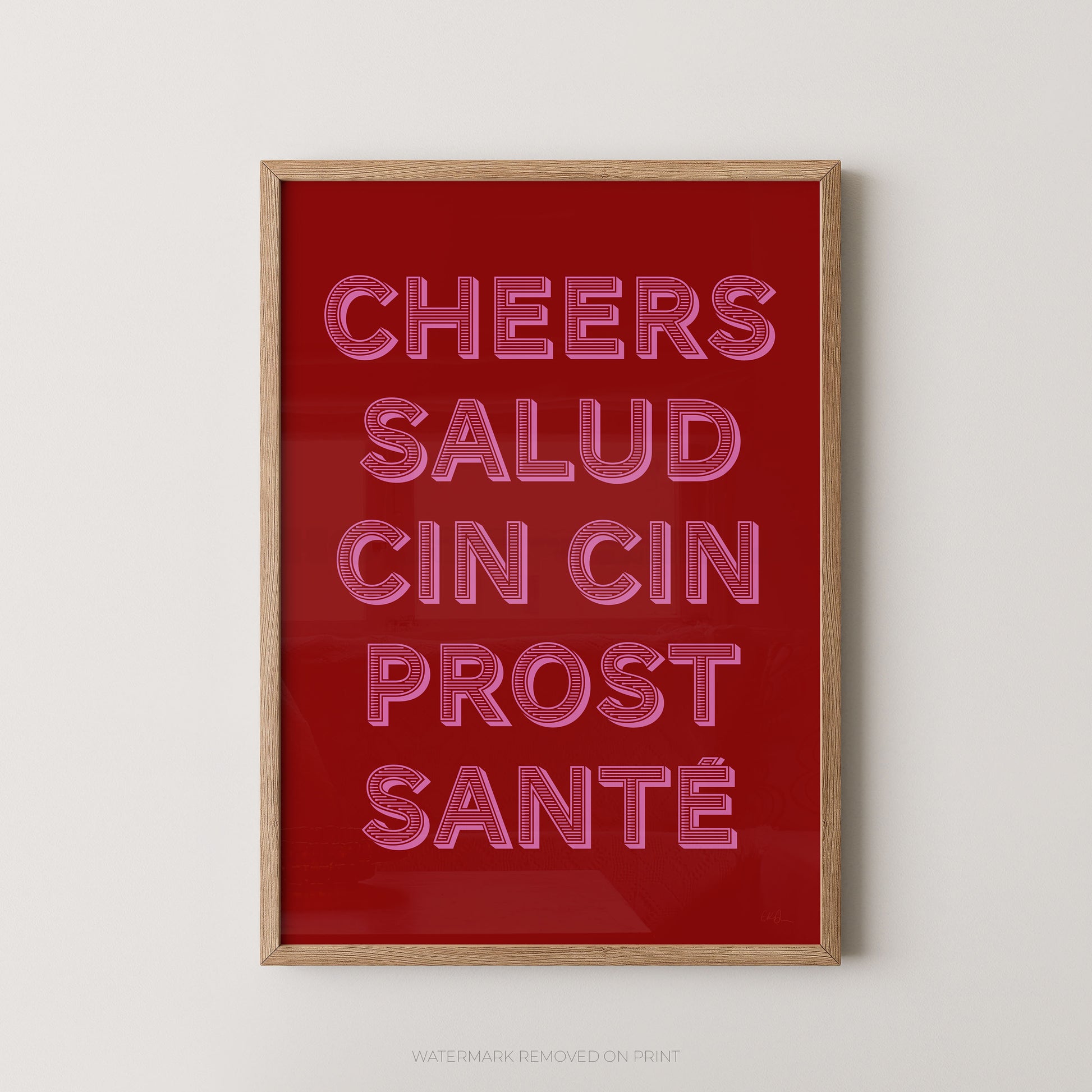 Framed wall art print with the phrase 'CHEERS SALUD CIN CIN PROST SANTÉ' in pink neon-style outlined lettering on a deep red background, shown in a light wood frame against a light gray wall.