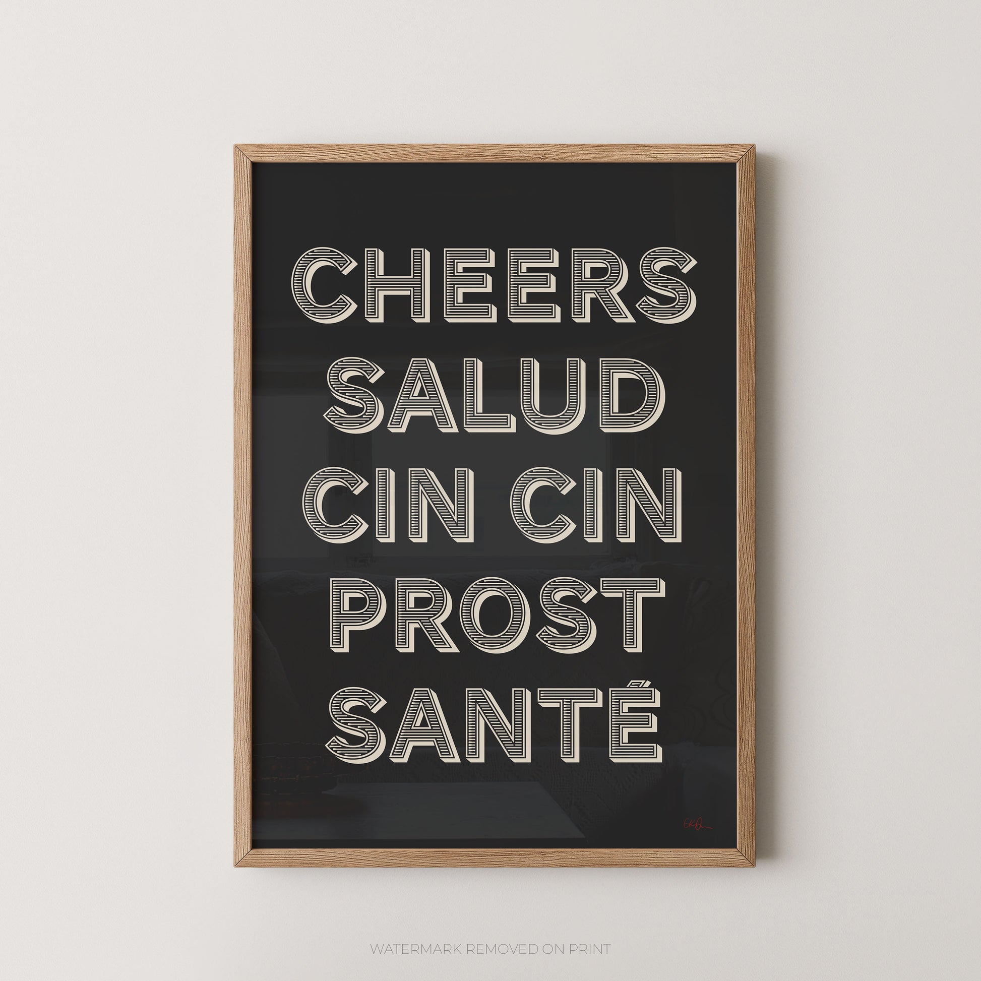 Framed wall art print with the phrase 'CHEERS SALUD CIN CIN PROST SANTÉ' in large metallic-style letters on a dark background, shown in a light wood frame against a plain light wall.