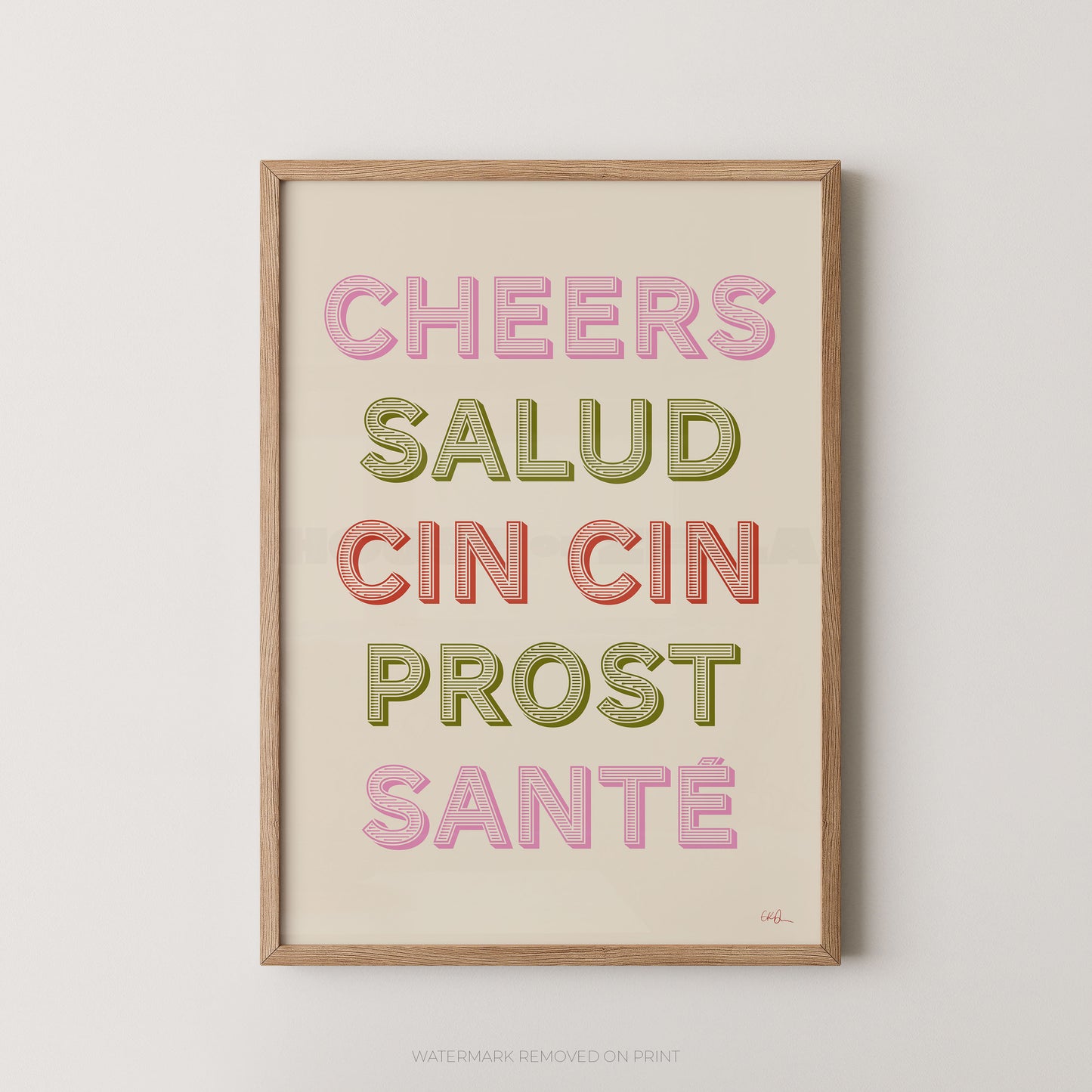 A framed typographic wall art print with the phrase “CHEERS SALUD CIN CIN PROST SANTÉ” in pastel pink, olive green, and coral lettering on a cream background, shown against a light neutral wall.