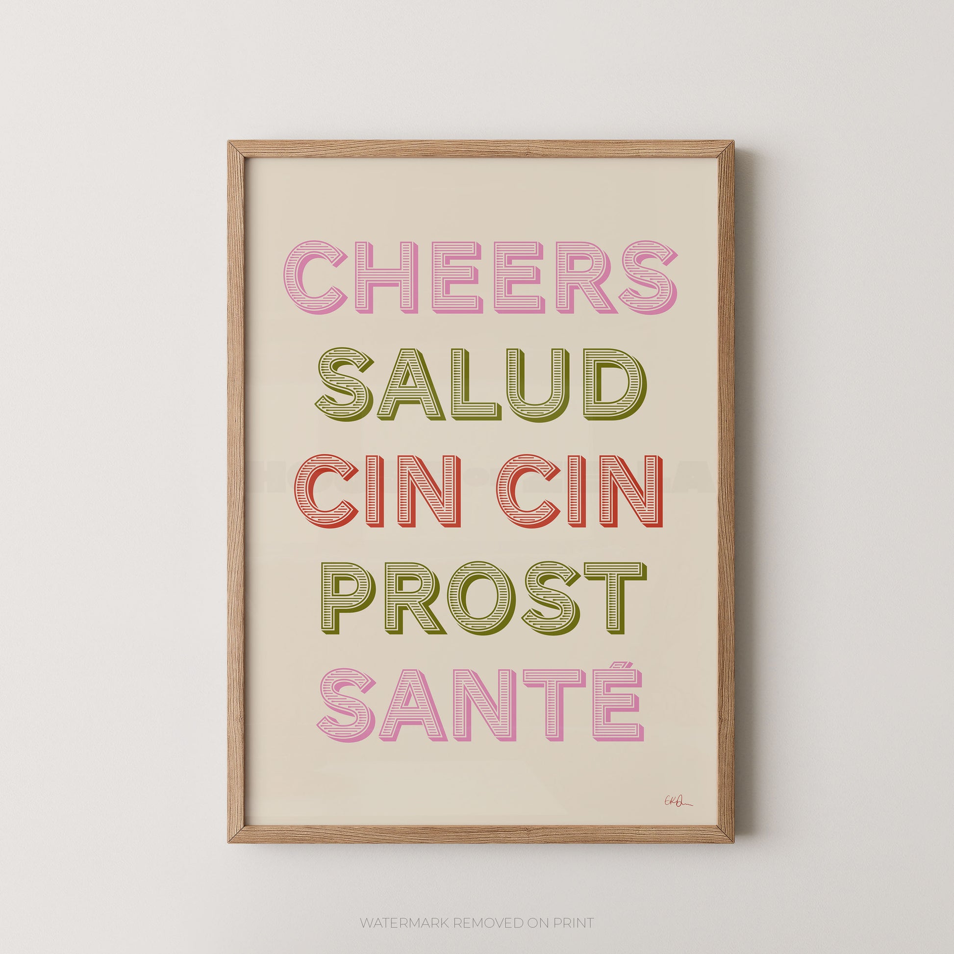 A framed typographic wall art print with the phrase “CHEERS SALUD CIN CIN PROST SANTÉ” in pastel pink, olive green, and coral lettering on a cream background, shown against a light neutral wall.